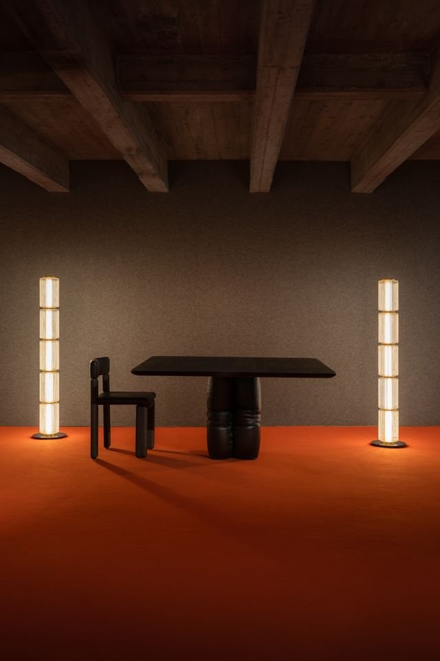 Une pièce minimaliste avec un plafond en bois, un mur gris clair, un tapis rouge, une table noire, une petite chaise noire, et deux lampes verticales allumées, positionnées de chaque côté de la table.