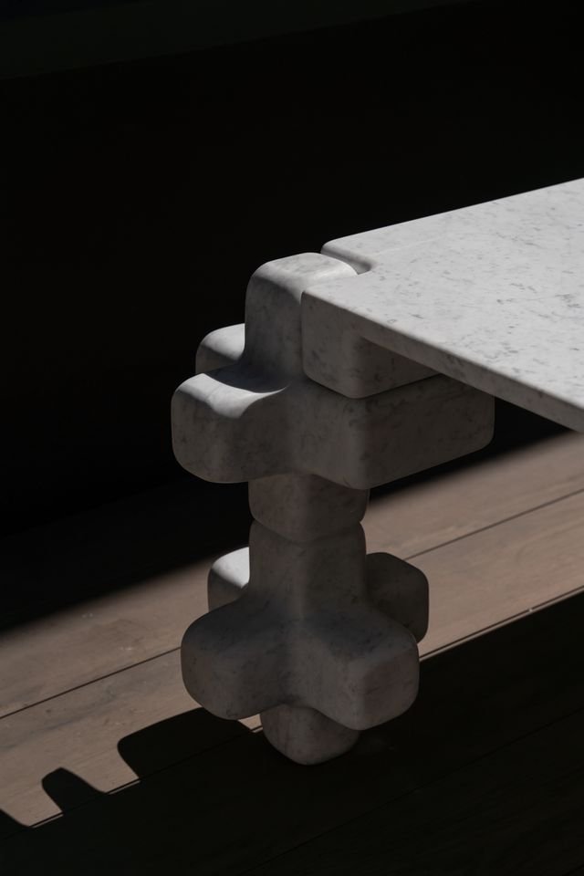 Table en marbre blanc avec un pied en forme de sculpture géométrique, placé sur un plan en bois, avec un fond sombre.