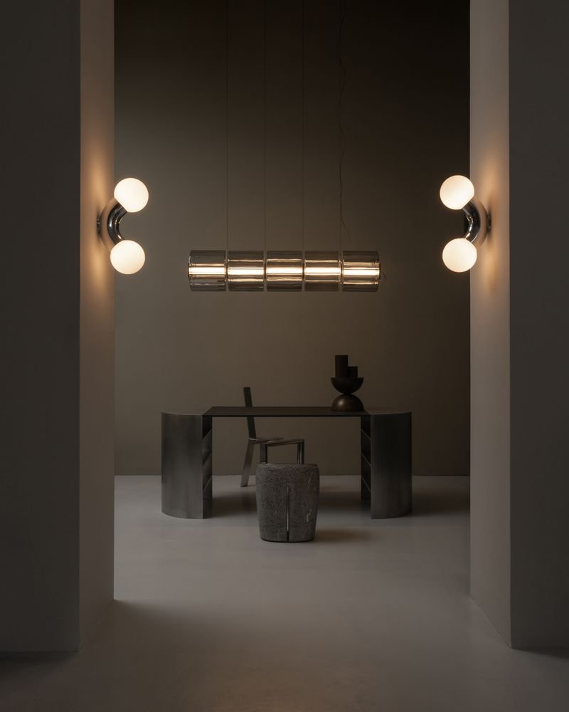 Vue d'un espace minimaliste avec un bureau moderne, deux chaises, une sculpture et un remarquable luminaire suspendu, encadré par deux murs avec appliques lumineuses.