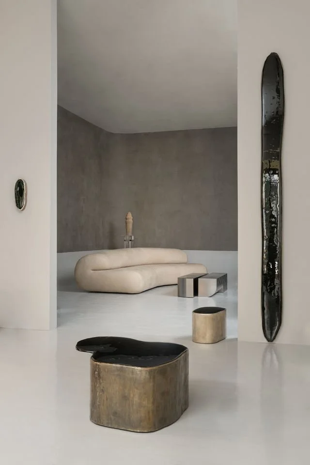 Une pièce moderne avec un canapé beige en forme de vague, des petits meubles en métal, une sculpture en bois sur un mur gris, et des objets décoratifs en bois et en céramique dans un style minimaliste.
