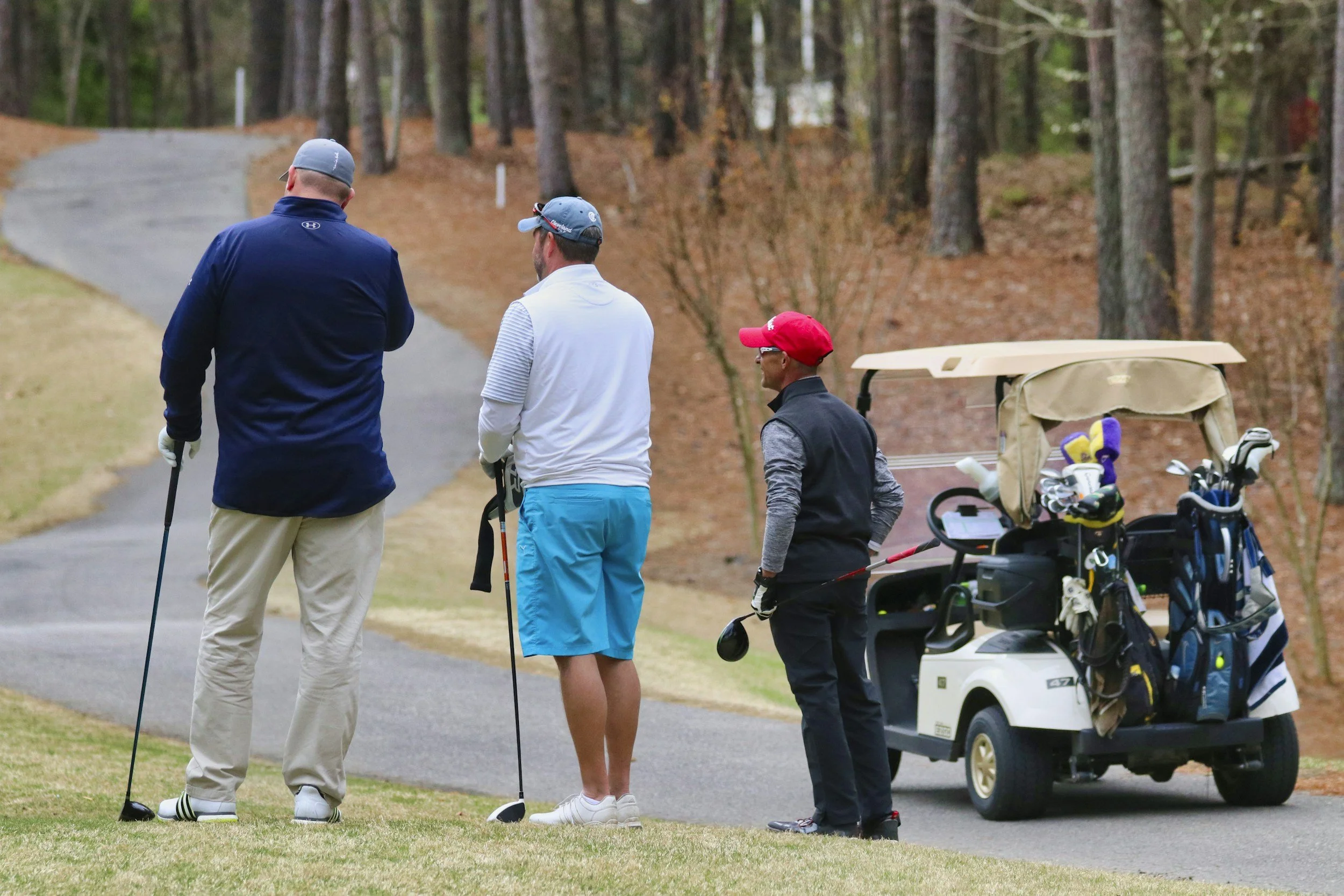 Three men on a golf course talking, with a golf cart carrying golf clubs nearby, in a wooded area.