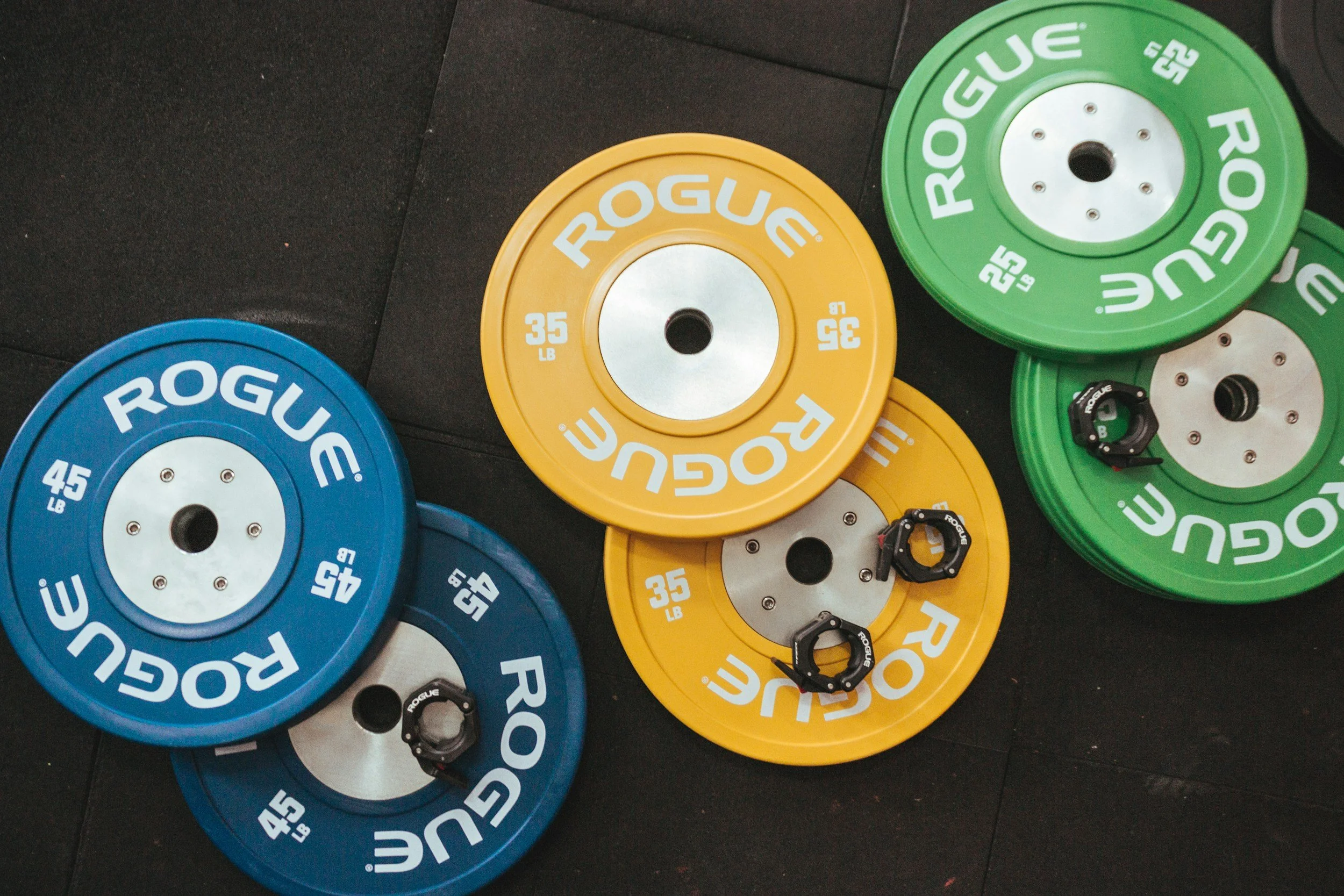 Colorful weightlifting bumper plates in yellow, blue, and green with Rogue branding, stacked and scattered on a black gym floor.