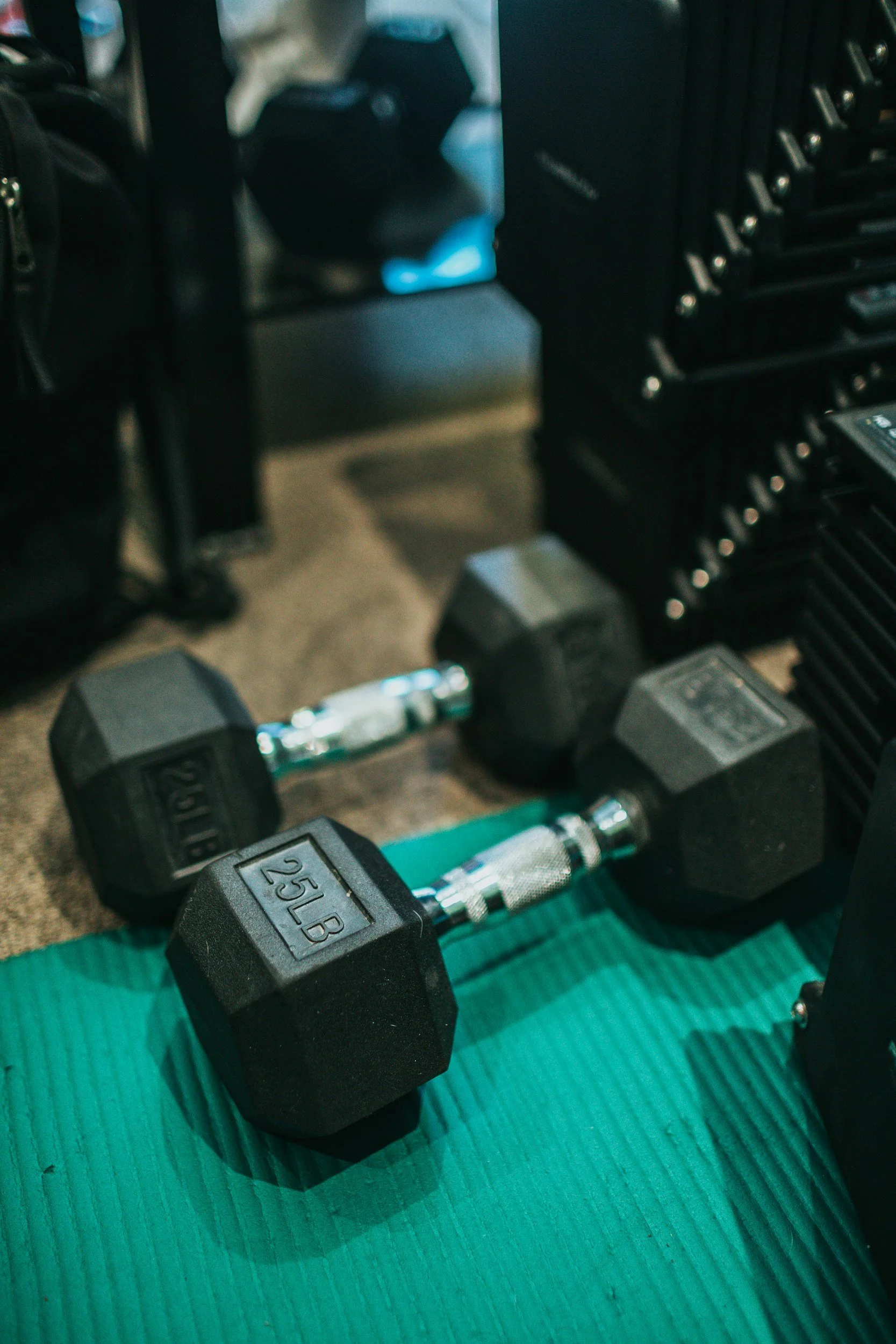 Pair of black 25-pound dumbbells on a teal exercise mat in a gym setting.