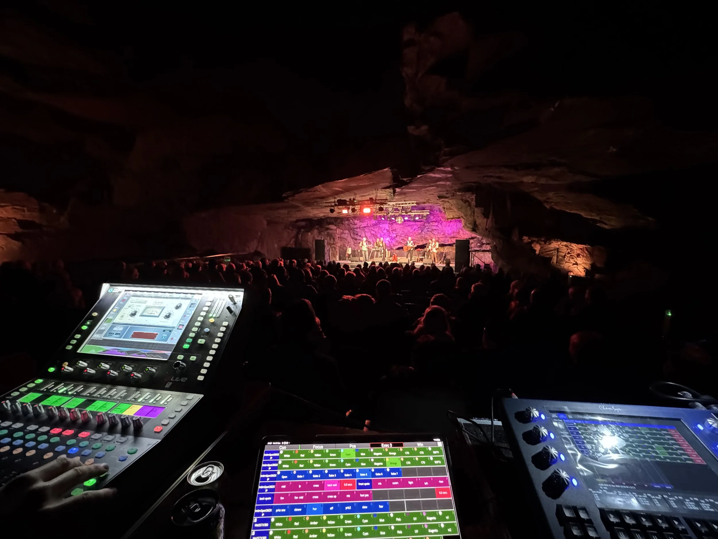 A concert is happening in a cave with a band playing on stage illuminated with pink and purple lights. The audience is seated in front of the stage. In the foreground, there are sound mixing consoles and equipment operated by a technician.
