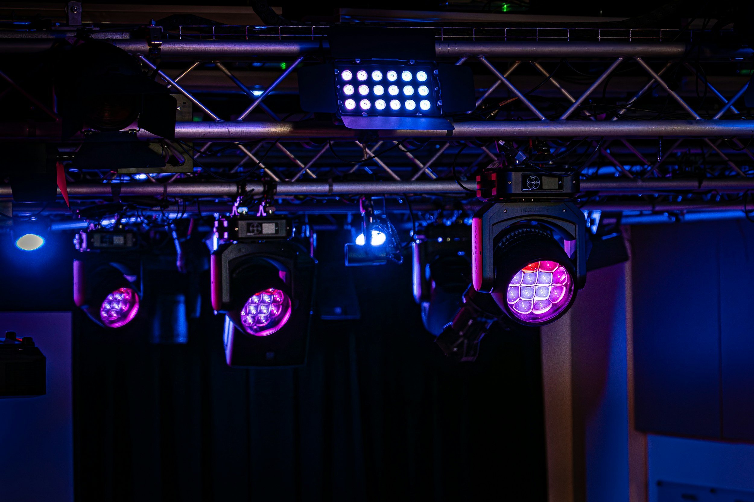 Stage lighting equipment with purple and blue LED lights and spotlights hanging from a metal truss in a dark room.
