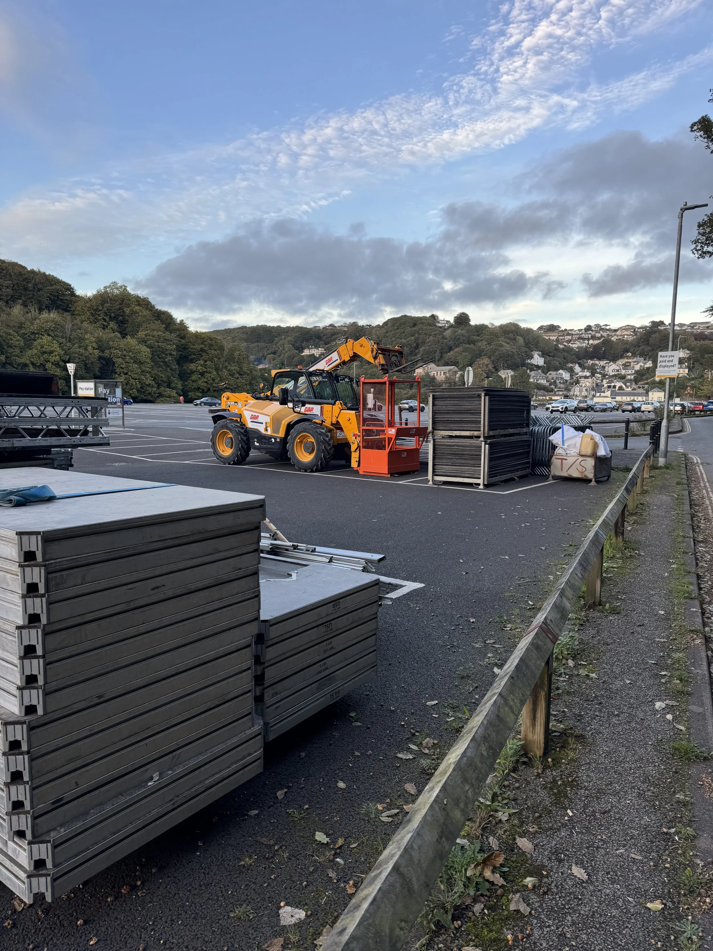 A parking lot with construction equipment, including a yellow loader with a front bucket and a lifting arm, surrounded by stacked black frames, a large plastic bag with a sign reading 'T/S', and a partially assembled table, against a backdrop of hills and cloudy sky.
