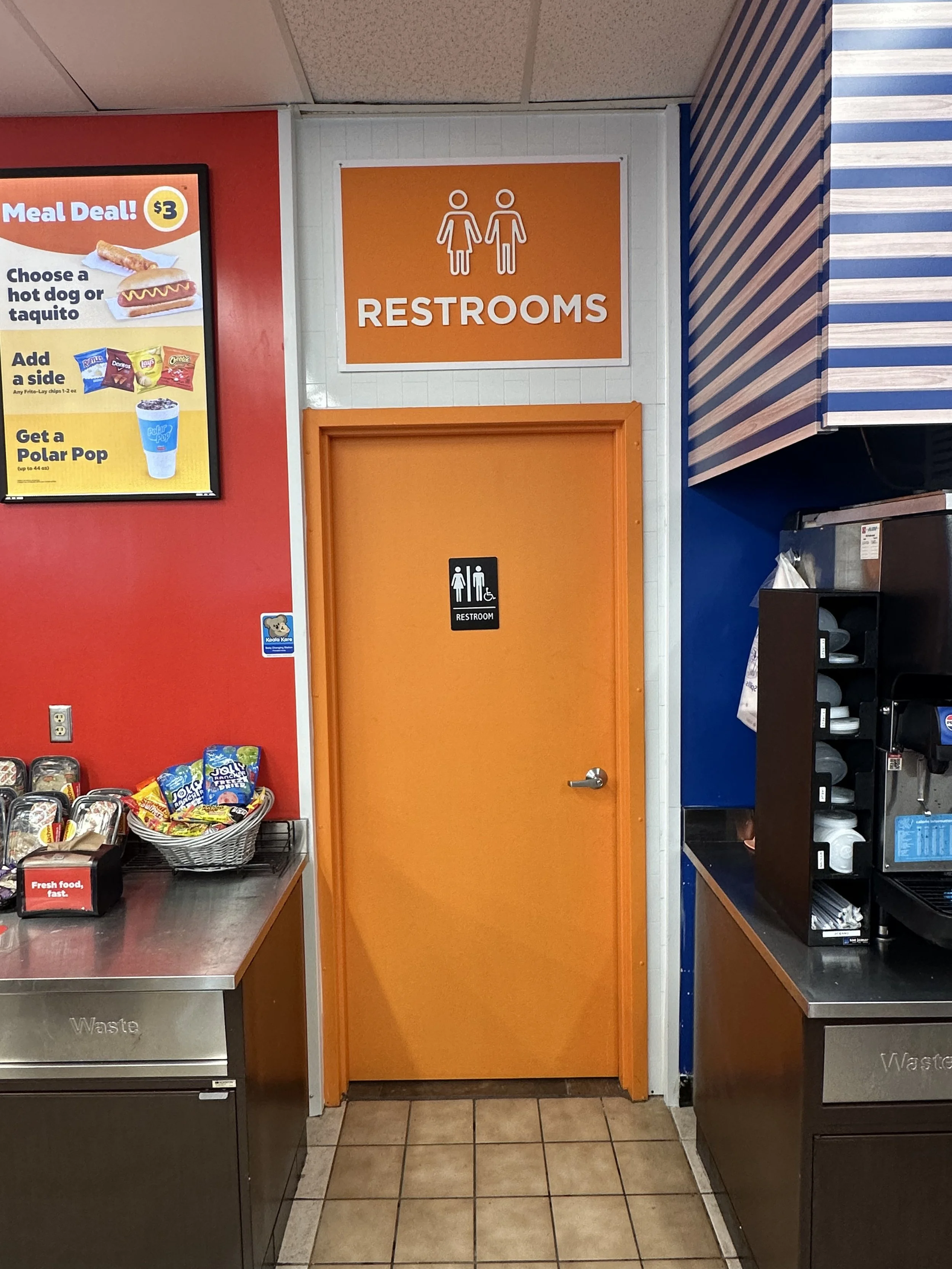 orange bathroom door with "restrooms" sign above, gender neutral, "meal deal" sign and gas station food outside the space