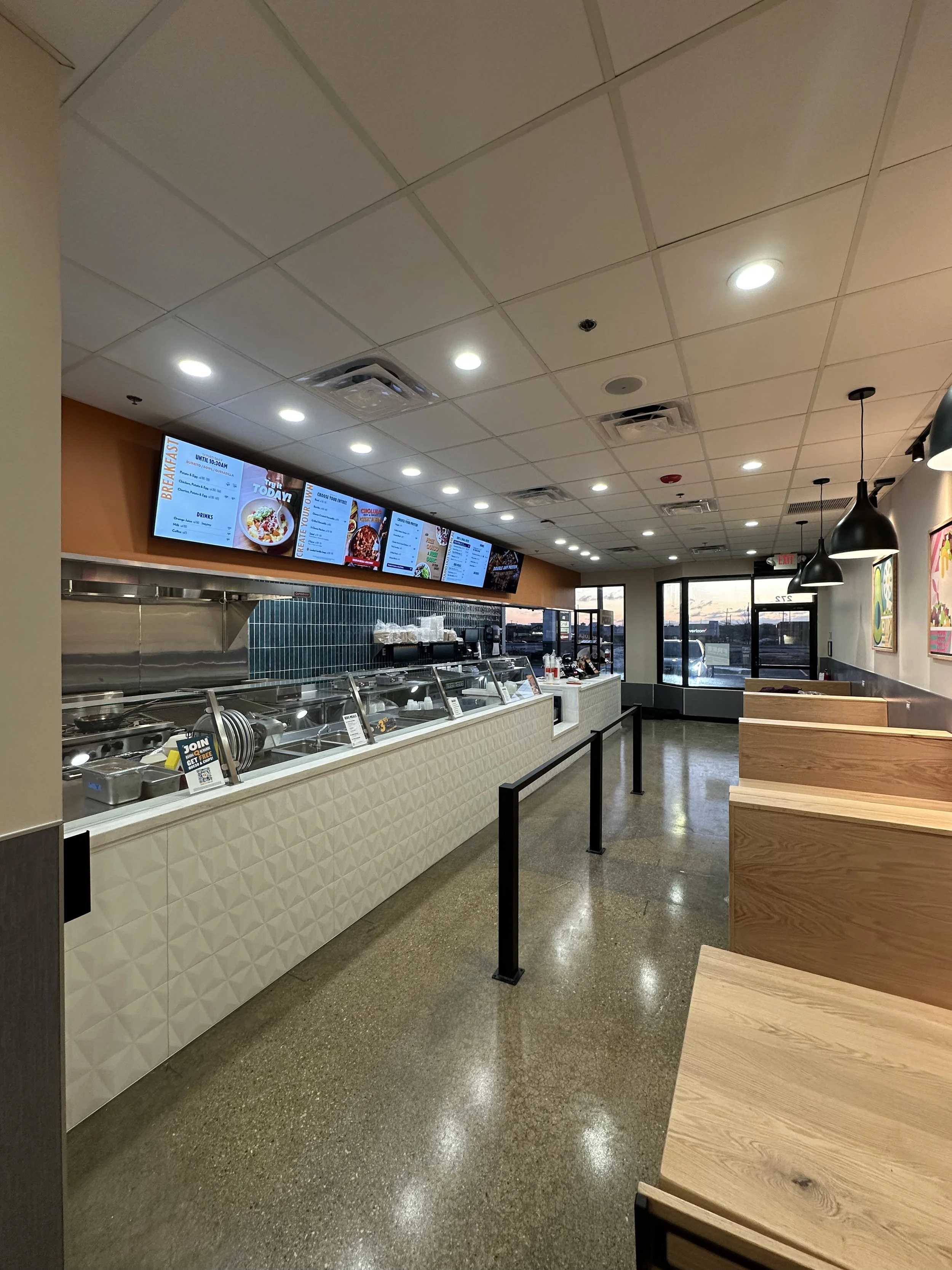 Qdoba restaurant counter with digital menus above, wooden booths with hanging pendants across from the counter