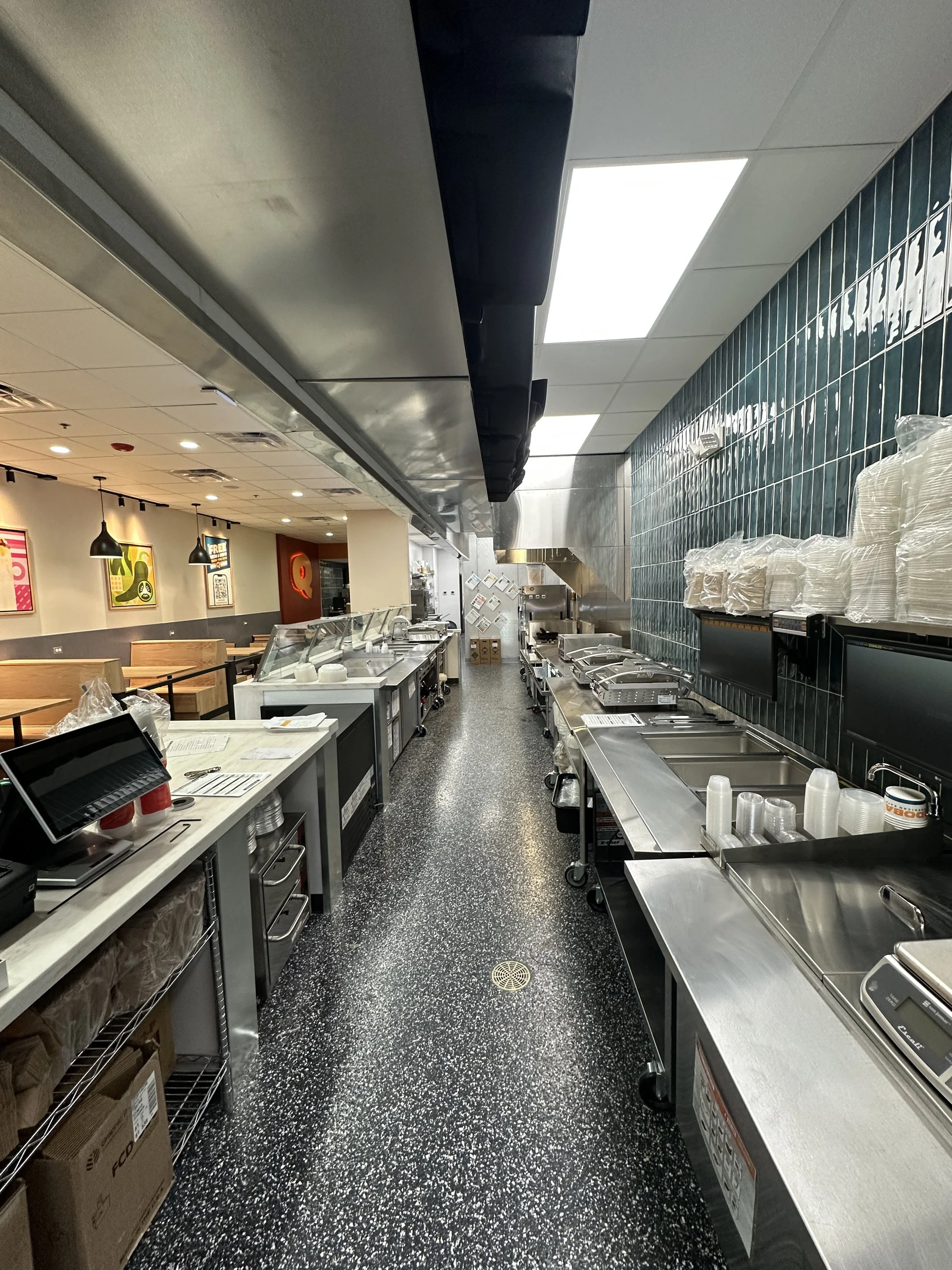 behind Qdoba restaurant counter, food selection counter on the left, food prep and sinks on the right, dark floor, bright lights