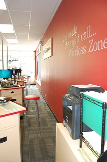 red wall in verizon store, gray carpet, light wood consoles on the left for setting up accounts