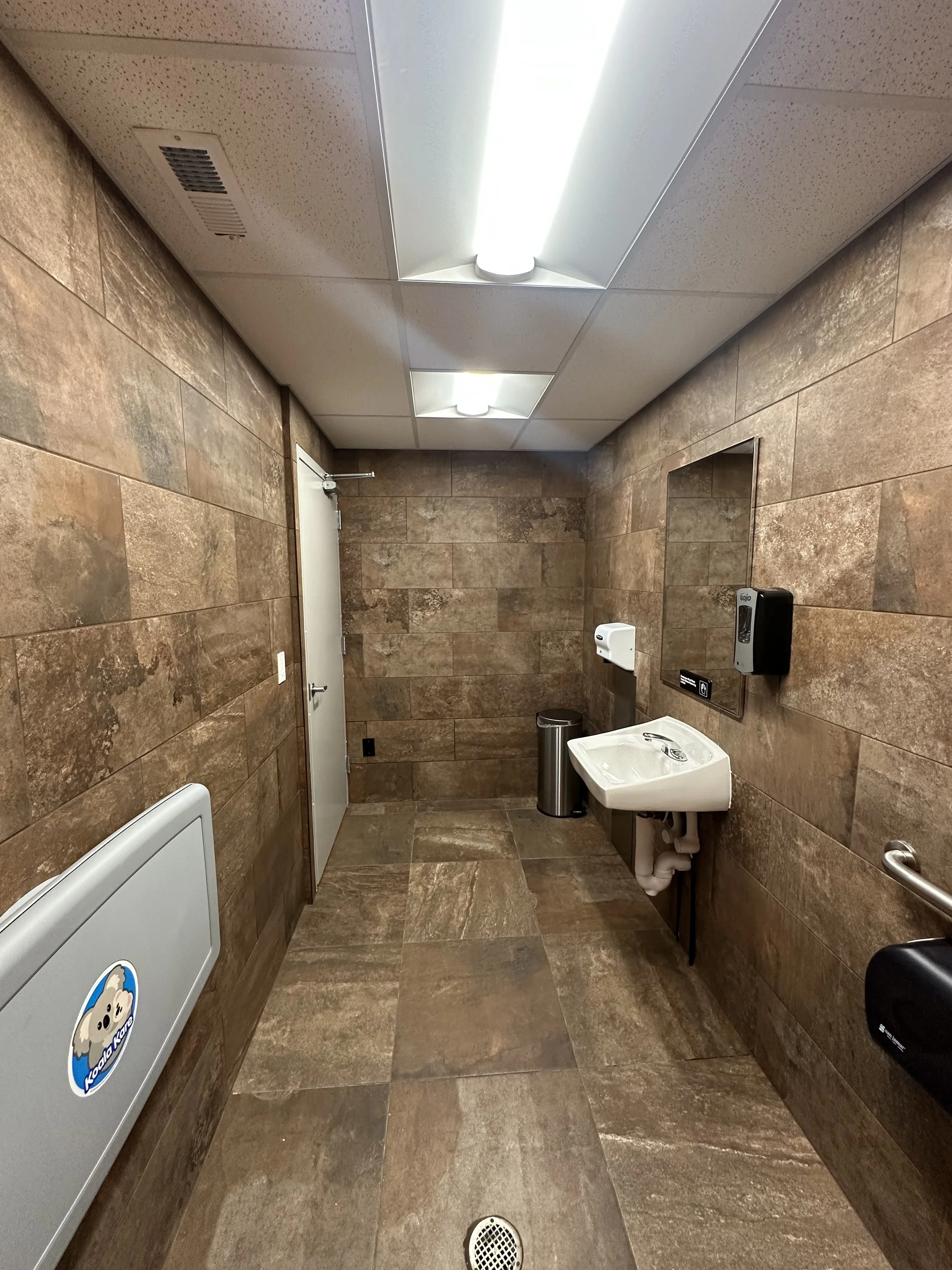 brown tile on floor and walls, flourescent lighting, bathroom sink, mirror, and door