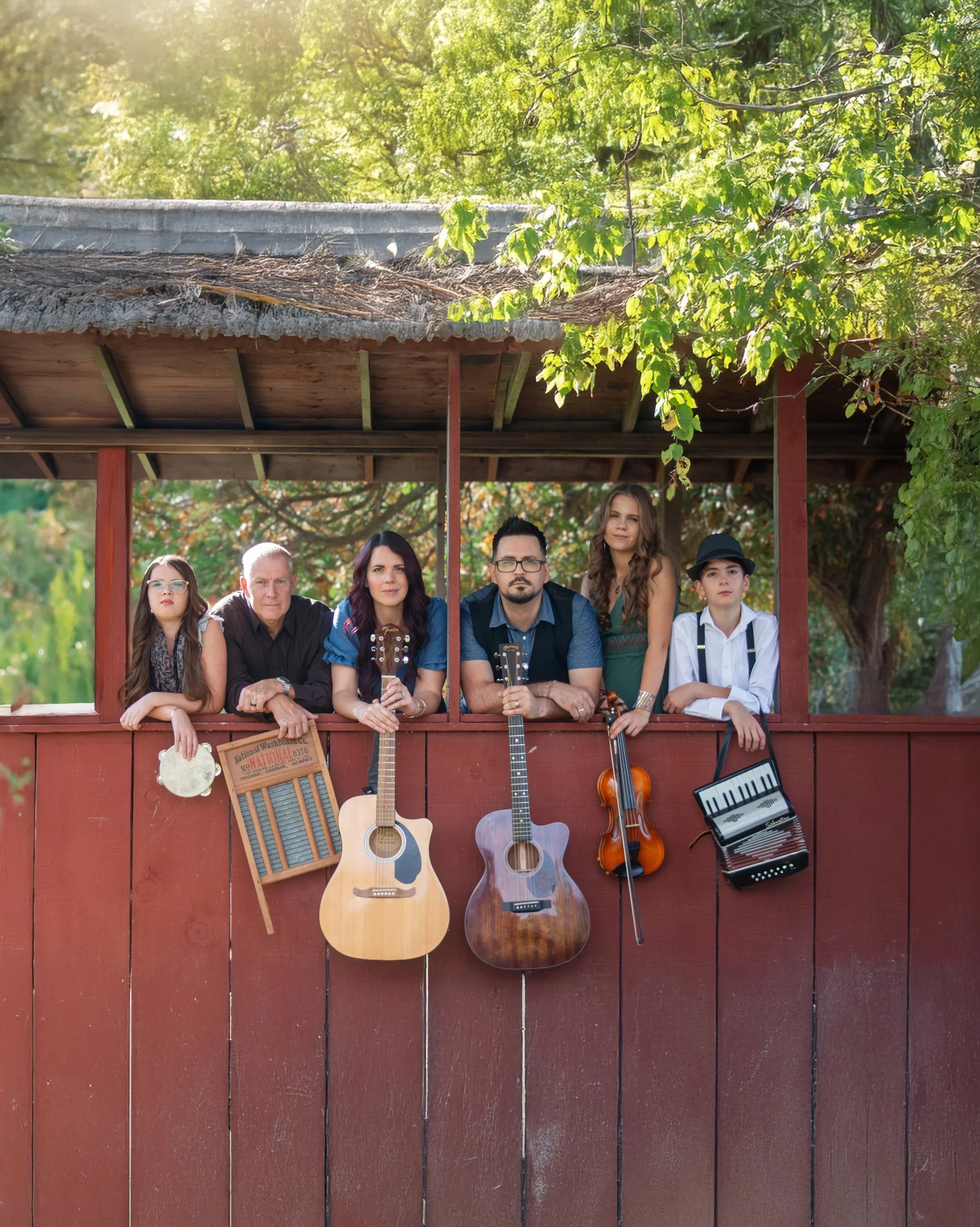 Six people holding musical instruments leaning on a red wooden fence in a park or garden with trees and green foliage in the background.