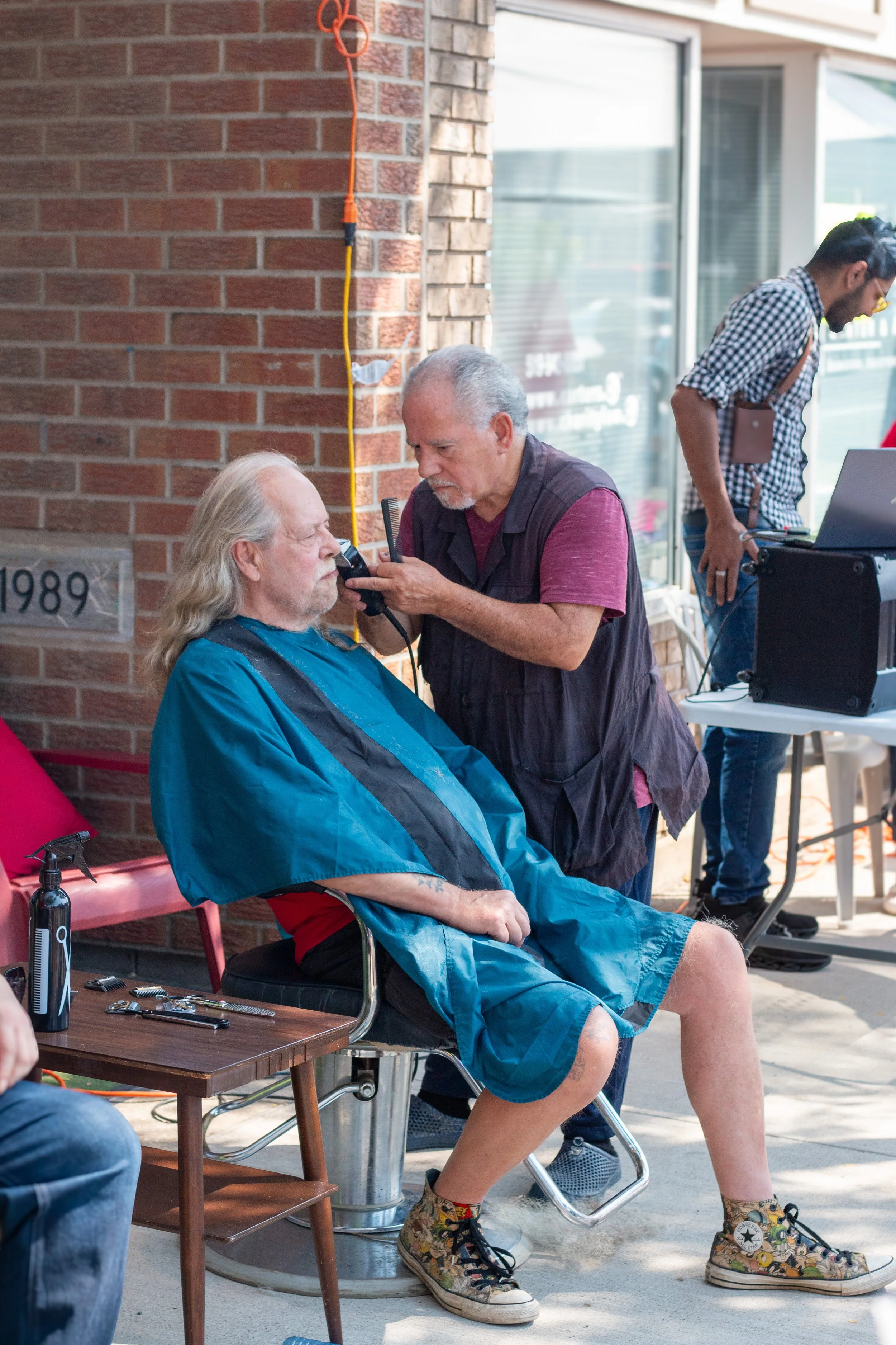 An elderly man with long gray hair and a beard sits in a barber chair with a blue cape around him, getting a beard trim from a middle-aged man inside a salon or barbershop. A young man in the background appears to be working with audio equipment.