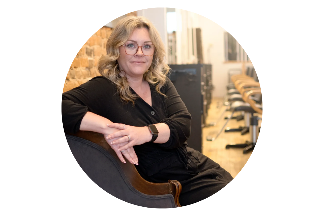 A woman with glasses and blonde hair, wearing a black blouse with rolled-up sleeves, sitting on a dark armchair in a room with exposed brick wall and wooden flooring, shelves, and chairs in the background.