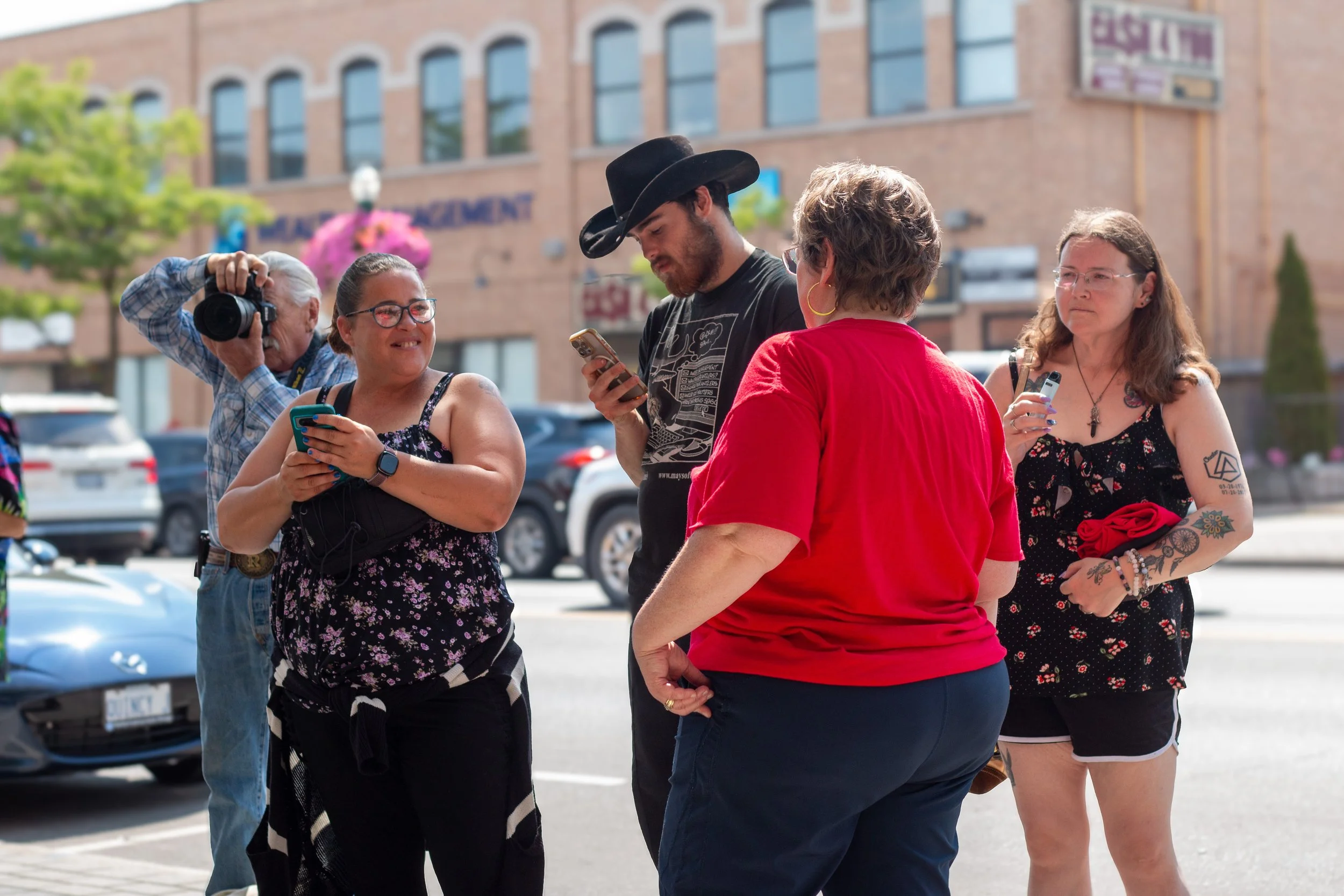 A group of five people standing in a parking lot in front of buildings, engaging in conversation and taking photos on their phones. One person is wearing a cowboy hat, and another has tattoos.