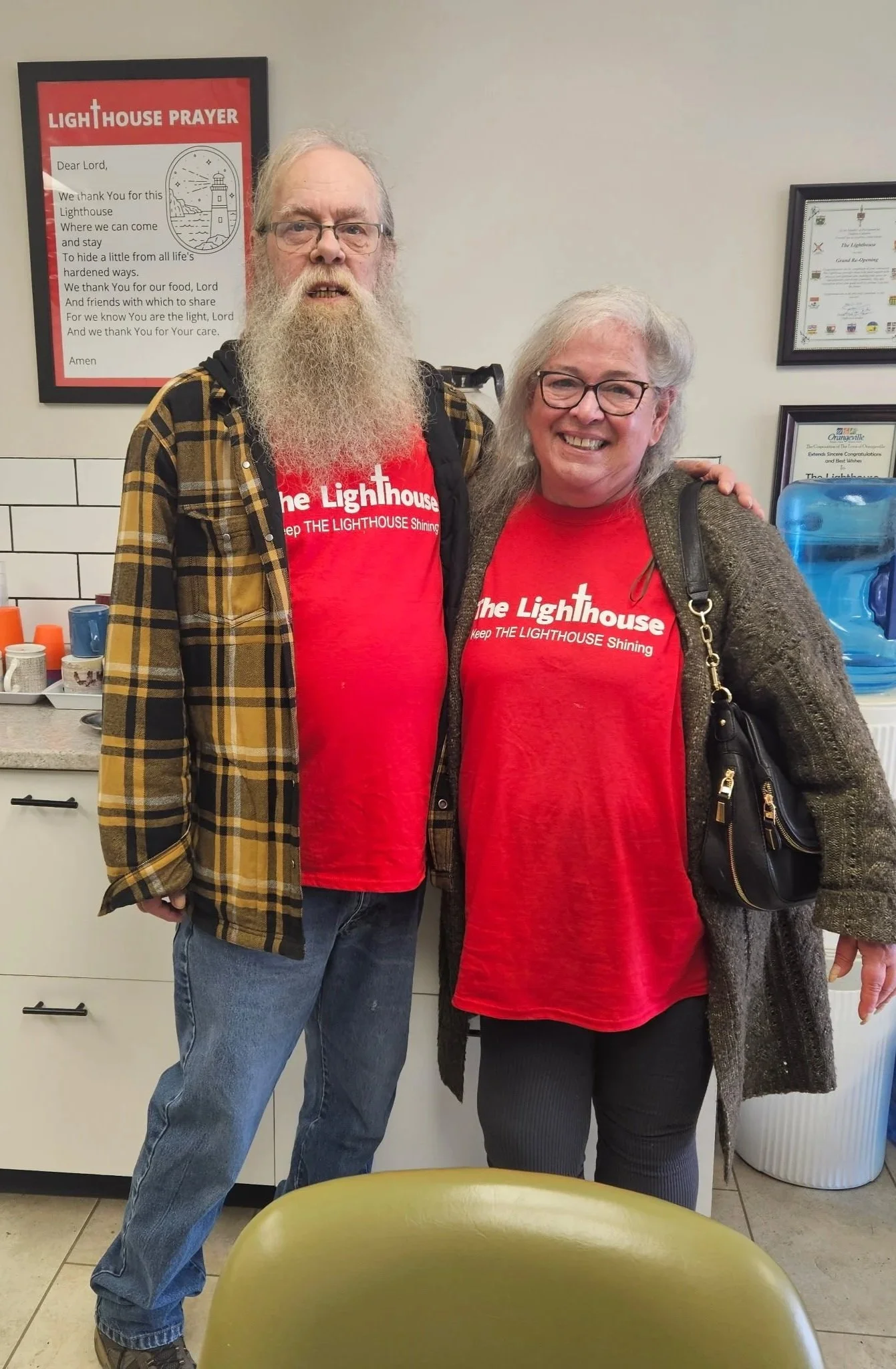 Two people standing together, both wearing red t-shirts that say 'The Lighthouse' with a lighthouse graphic and the phrase 'Keep the Lighthouse Shining.' They are indoors in a room with framed certificates and a poster that reads 'Lighthouse Prayer,' a water cooler, and a countertop with cups and a coffee pot. The person on the left has a long white beard, glasses, and is wearing a yellow, black, and white plaid shirt over the red t-shirt, while the person on the right has shoulder-length gray hair, glasses, and is wearing a gray cardigan over the red t-shirt. Both are smiling.