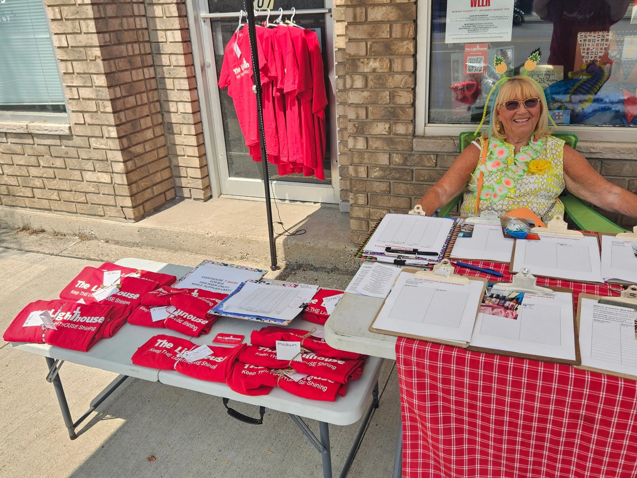 A smiling elderly woman with blonde hair wearing sunglasses and a colorful outfit sitting behind a table with red T-shirts, clipboards, and paperwork outside a brick building.