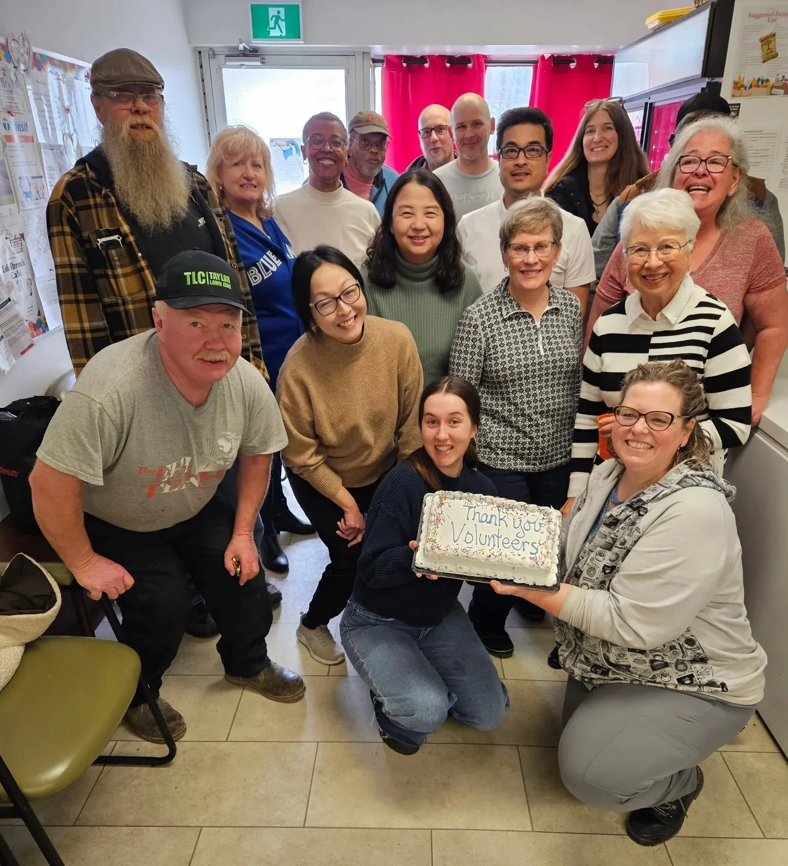 Group of diverse volunteers smiling, holding a cake that says 'Thank you Volunteers,' inside a community room.