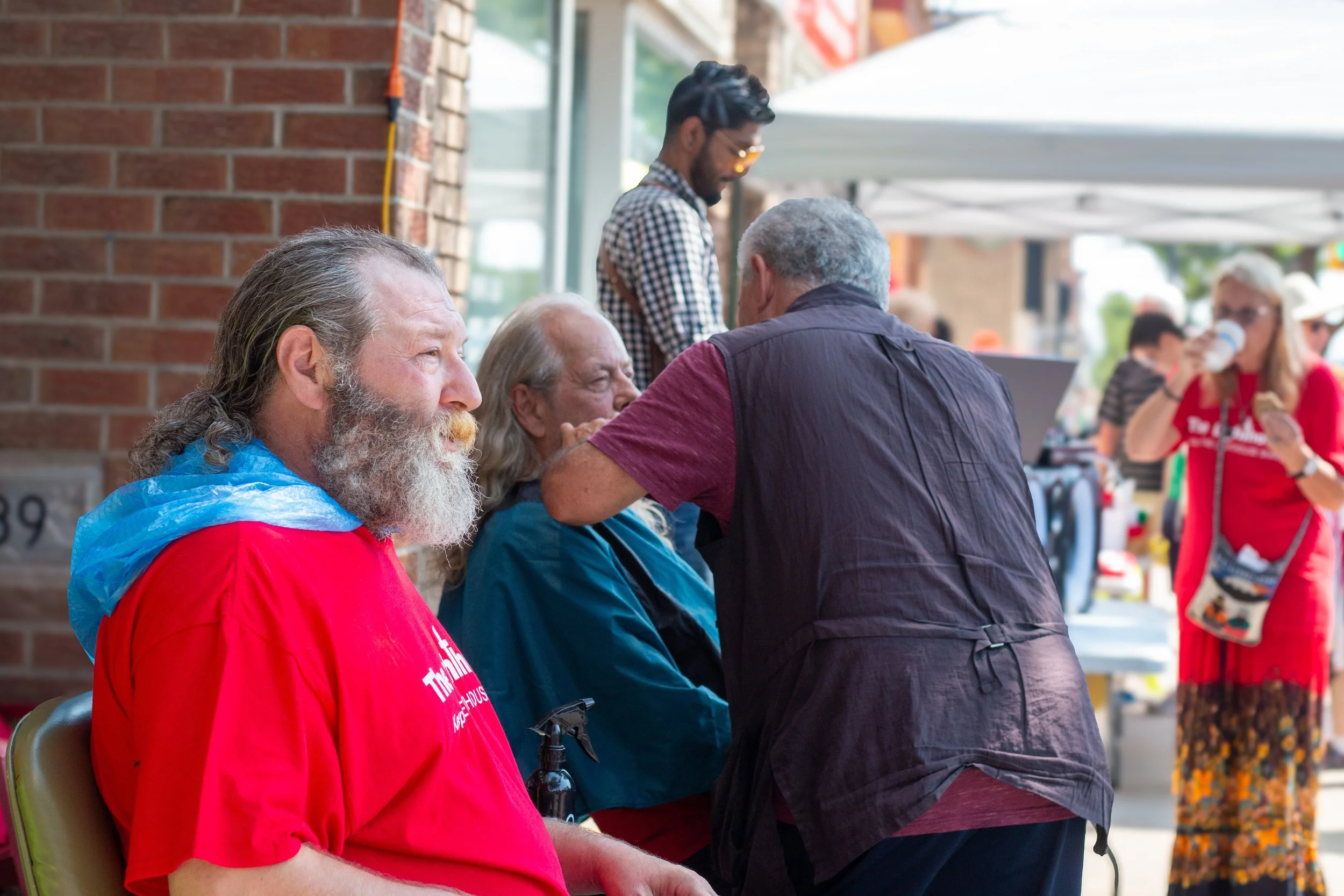 A man with a white beard sitting on a chair while another person is giving him a health checkup or applying a treatment to his face. People are socializing outdoors under tents, with some enjoying food and drinks.