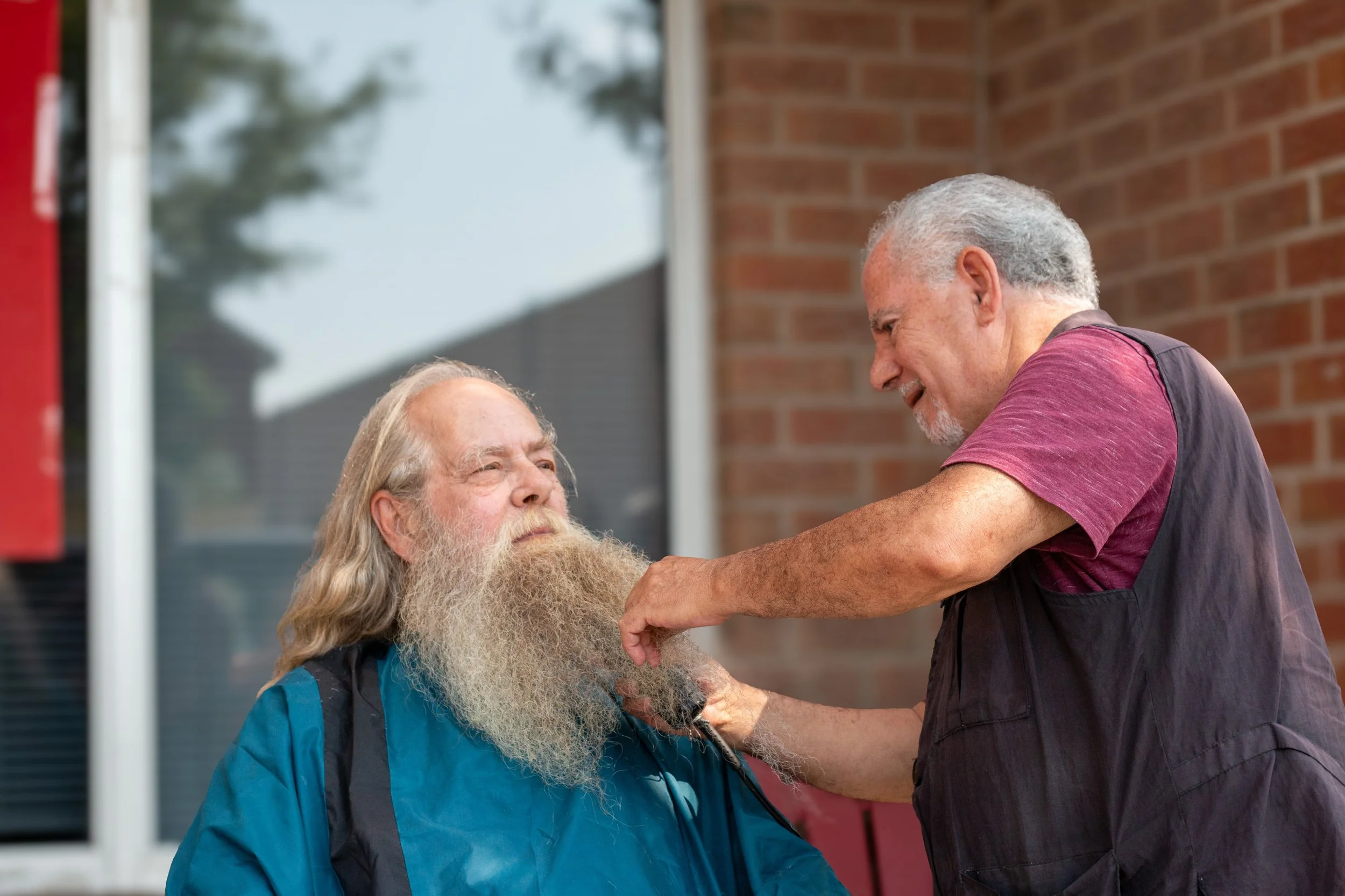 A man with a long beard sitting outside a building while another man with gray hair styling his beard.