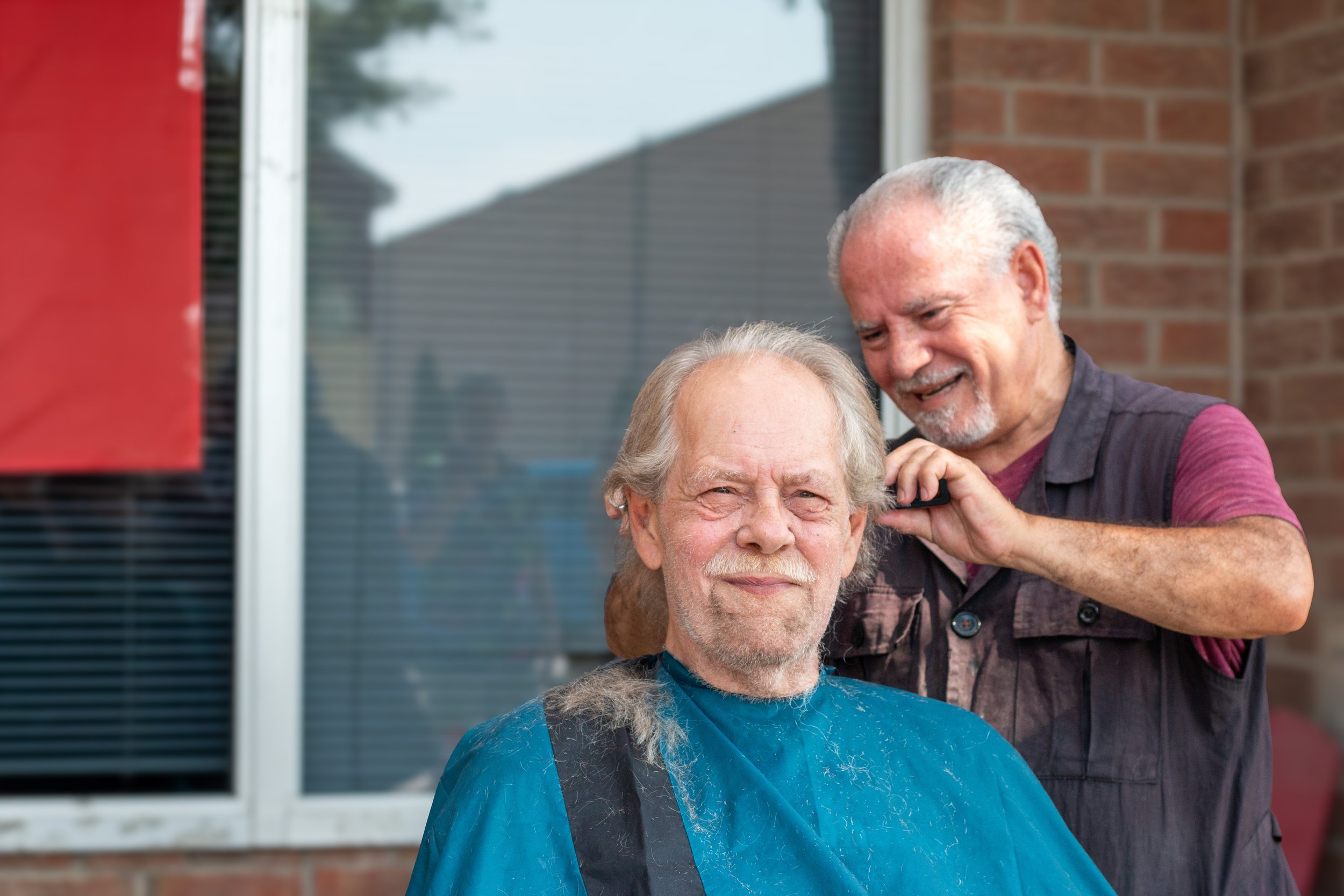 An elderly man with long gray hair and a beard getting a haircut outdoors at a barber shop, with a smiling barber standing behind him, both appearing content and happy.
