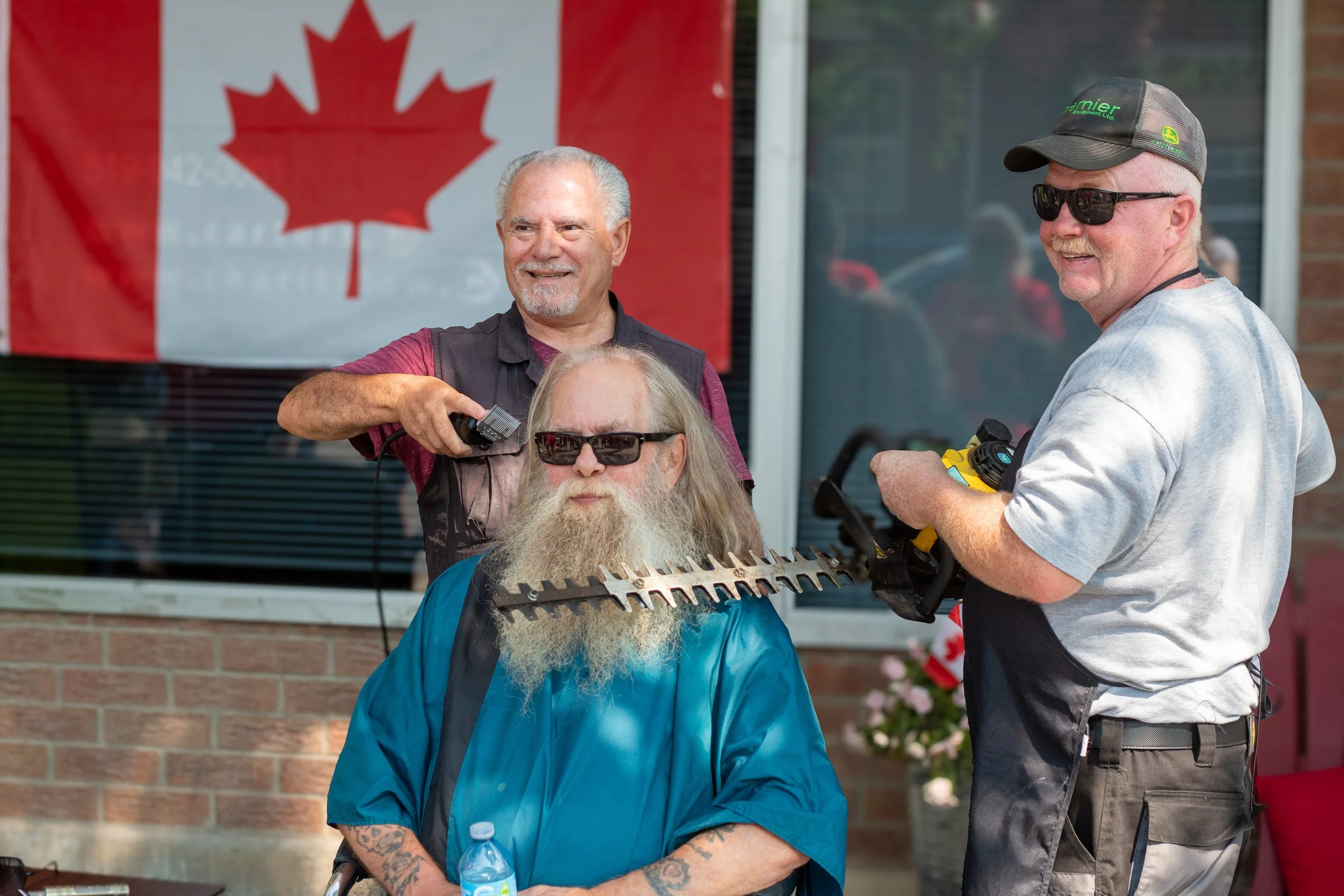 Three men, two standing and one seated, are outside a building in front of a Canadian flag. The seated man has a long beard, sunglasses, and is wearing a blue shirt. The man on the left is holding a hair clipper and smiling, and the man on the right 
