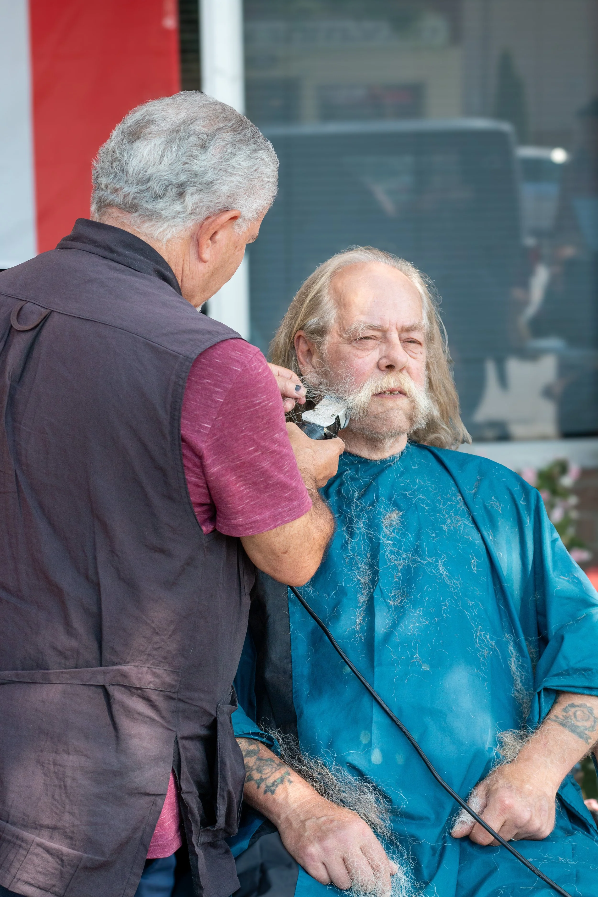 An older man with long gray hair and beard sitting outdoors, getting his beard trimmed by a barber.