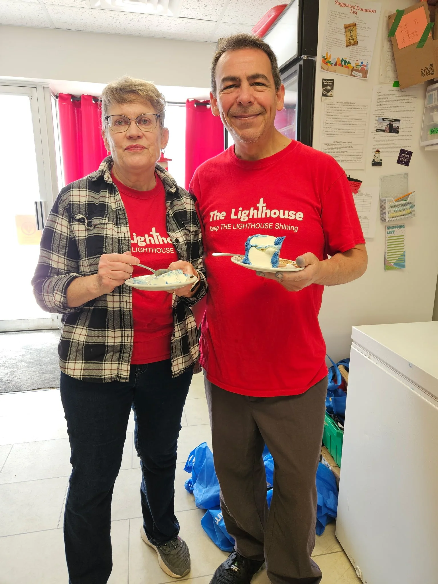 Two people, a woman and a man, stand inside a room holding plates of ice cream. They are smiling at the camera. Both are wearing red shirts with the logo and text 'The Lighthouse Keep THE LIGHTHOUSE Shining'. The woman has short gray hair, glasses, and is dressed in a plaid shirt over her red T-shirt. The man has short brown hair and a beard. The room has pink curtains, a black refrigerator, and various papers and signs on the wall.
