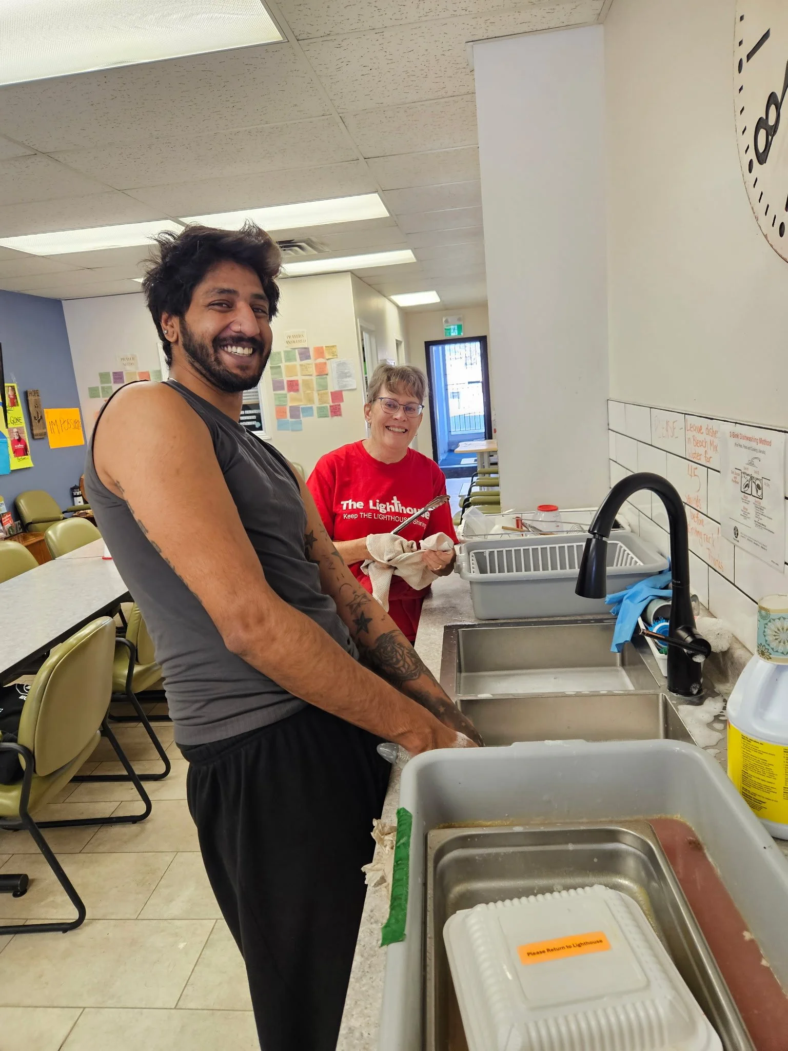 A man with dark hair and tattoos smiling at the camera, standing at a kitchen sink, with a woman in a red t-shirt behind him holding a paper towel, in a community kitchen or cafeteria setting.
