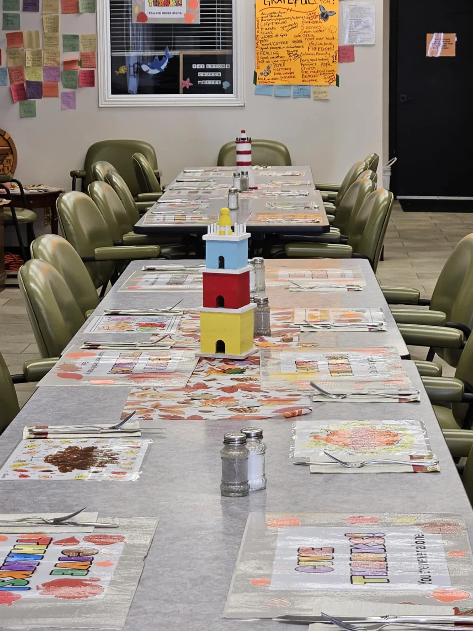 A long restaurant table set with colorful paper placemats, silverware, salt and pepper shakers, and small decorative lighthouse centerpieces. The background features a decorated wall with sticky notes, a large orange poster, and a window with paper whale and star decorations.
