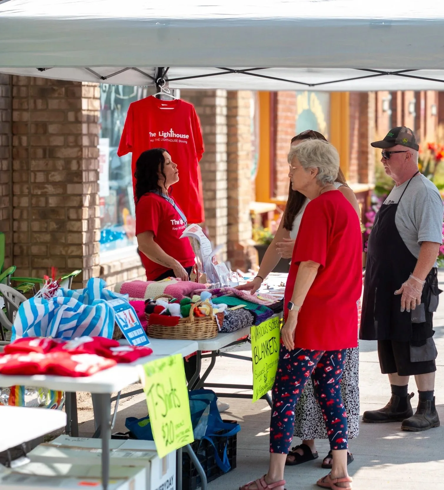 A street vendor booth selling various blankets and clothing items, with a red shirt hanging and several customers browsing and talking.