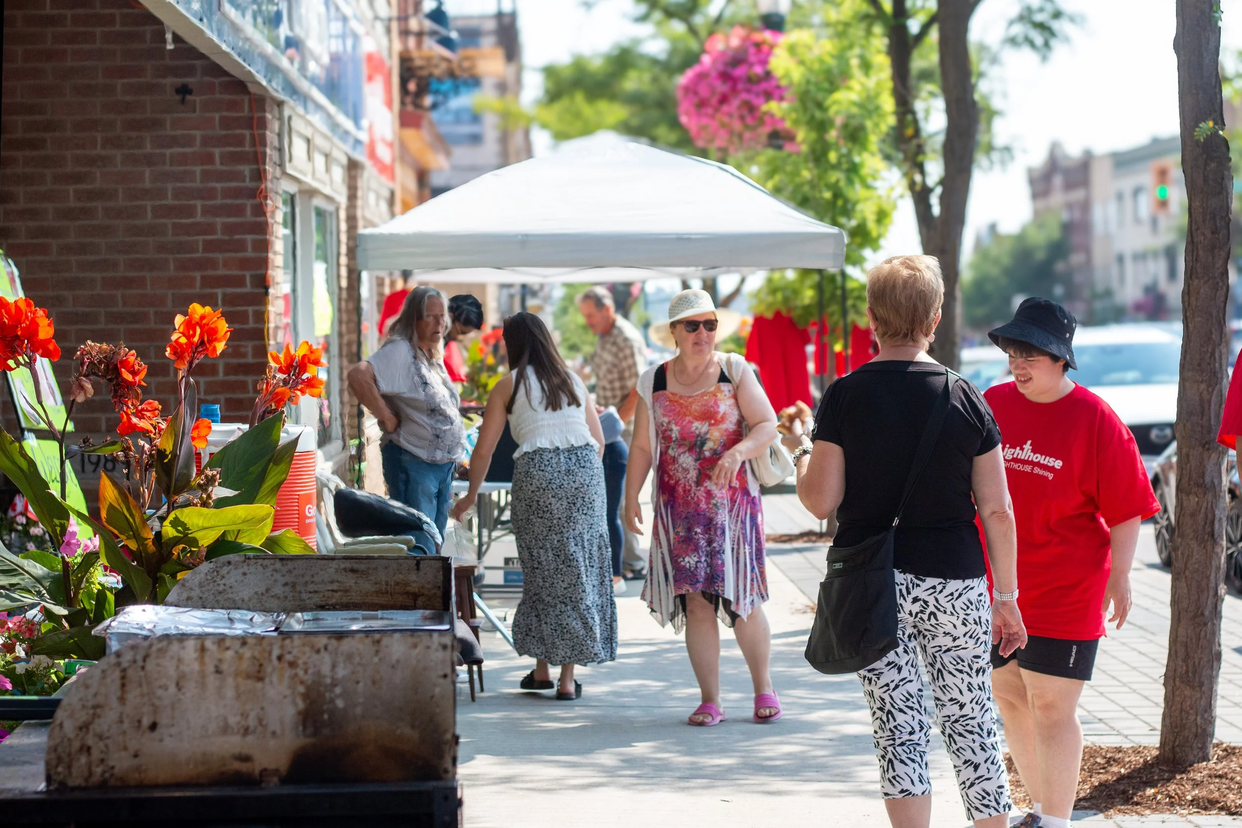 People browsing outdoor market stalls on a sunny day, with flowers and trees lining the sidewalk.