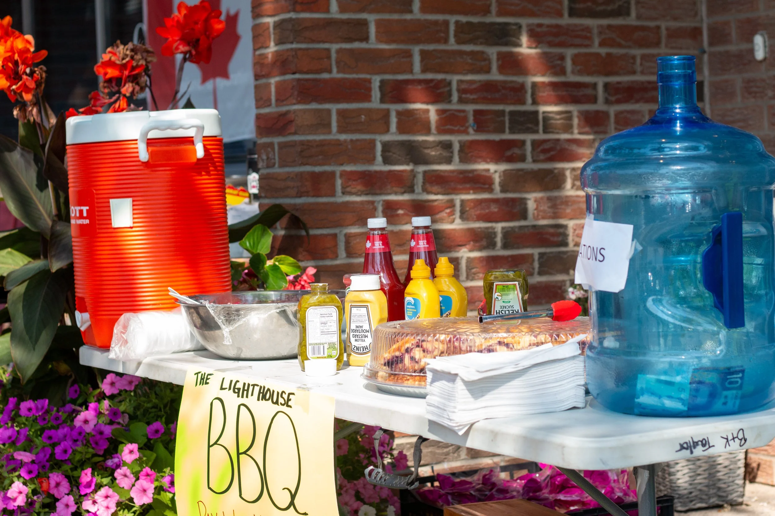 A table outdoors with a sign reading 'The Lighthouse BBQ,' containing condiments, pizza, drinks, paper towels, and a large water jug beside a brick wall and pink flowers.
