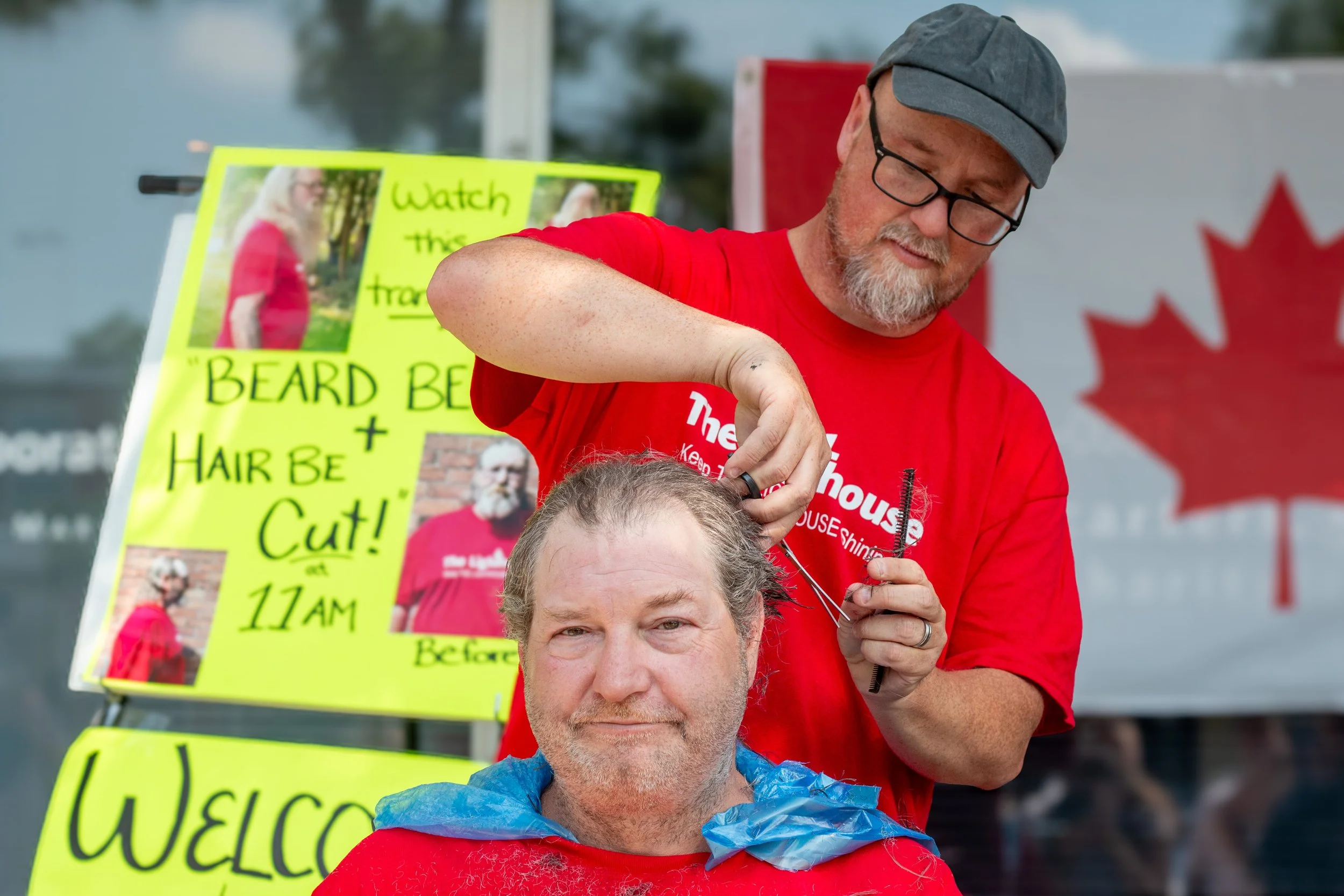 A man is giving a haircut to another man outdoors. The man receiving the haircut has gray hair and is wearing a red cape. The barber, wearing a red t-shirt, gray cap, and glasses, is trimming the gray hair. Behind them, there are colorful signs, one 