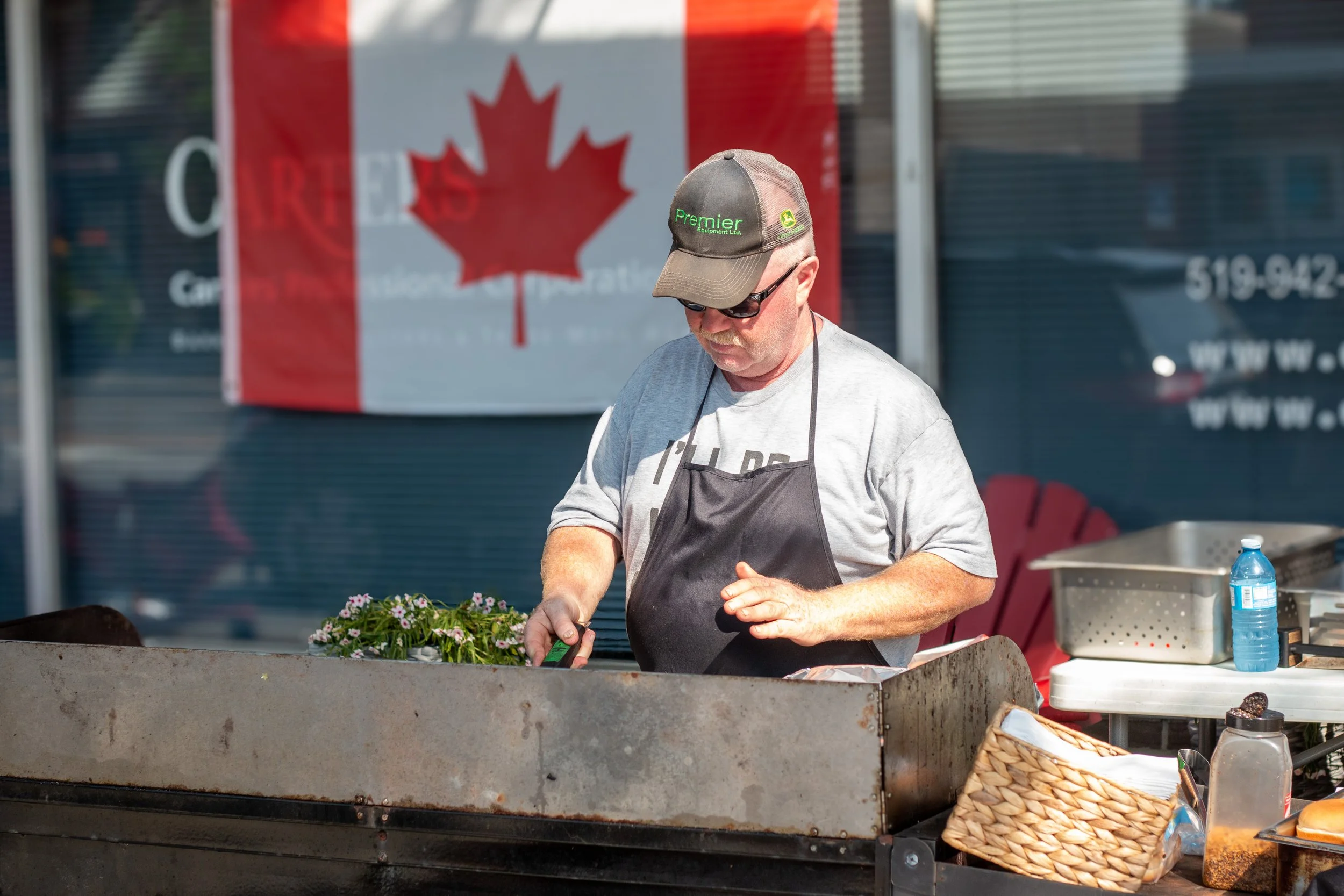 Man grilling outdoors, wearing a gray T-shirt, black apron, and a dark green baseball cap. Behind him, a Canadian flag is visible.