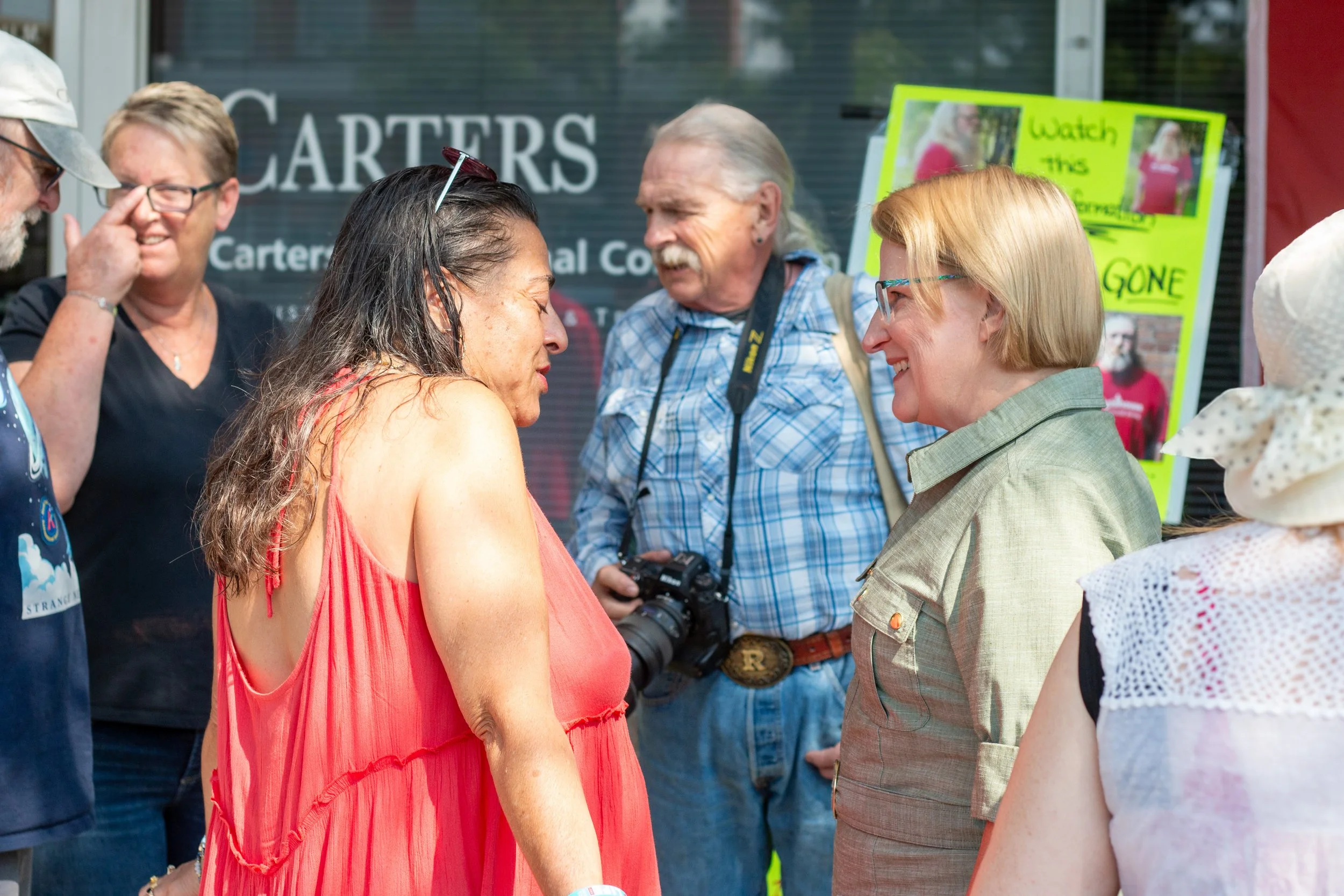 Group of people smiling and talking outdoors, with a sign in the background that reads 'Watch this' and 'GONE'.