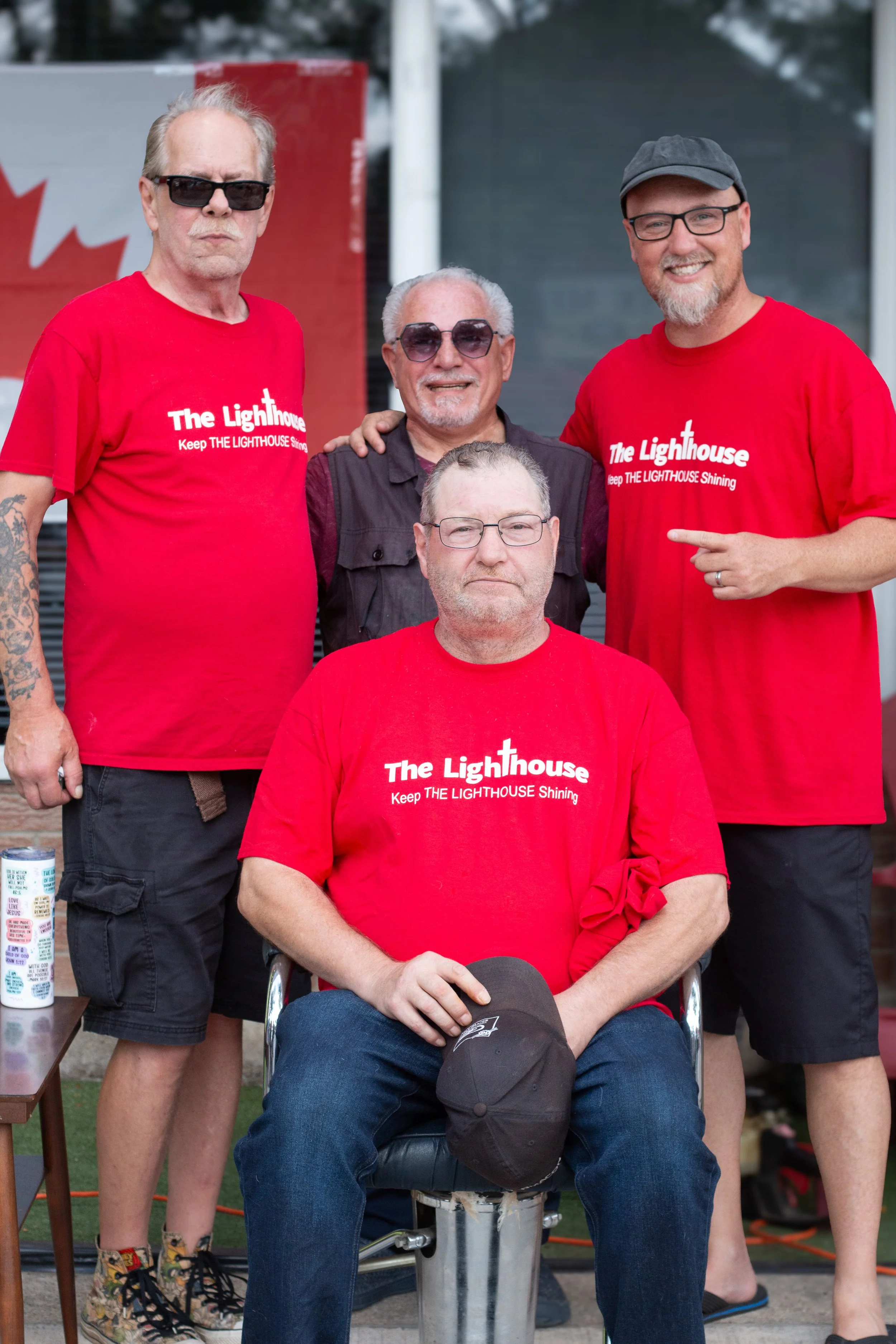 Four men posing together outdoors, three of them wearing matching red T-shirts with the phrase 'The Lighthouse Keep THE LIGHTHOUSE Shining' printed on them, and one man in a black vest. They are smiling and appear to be participating in a group event