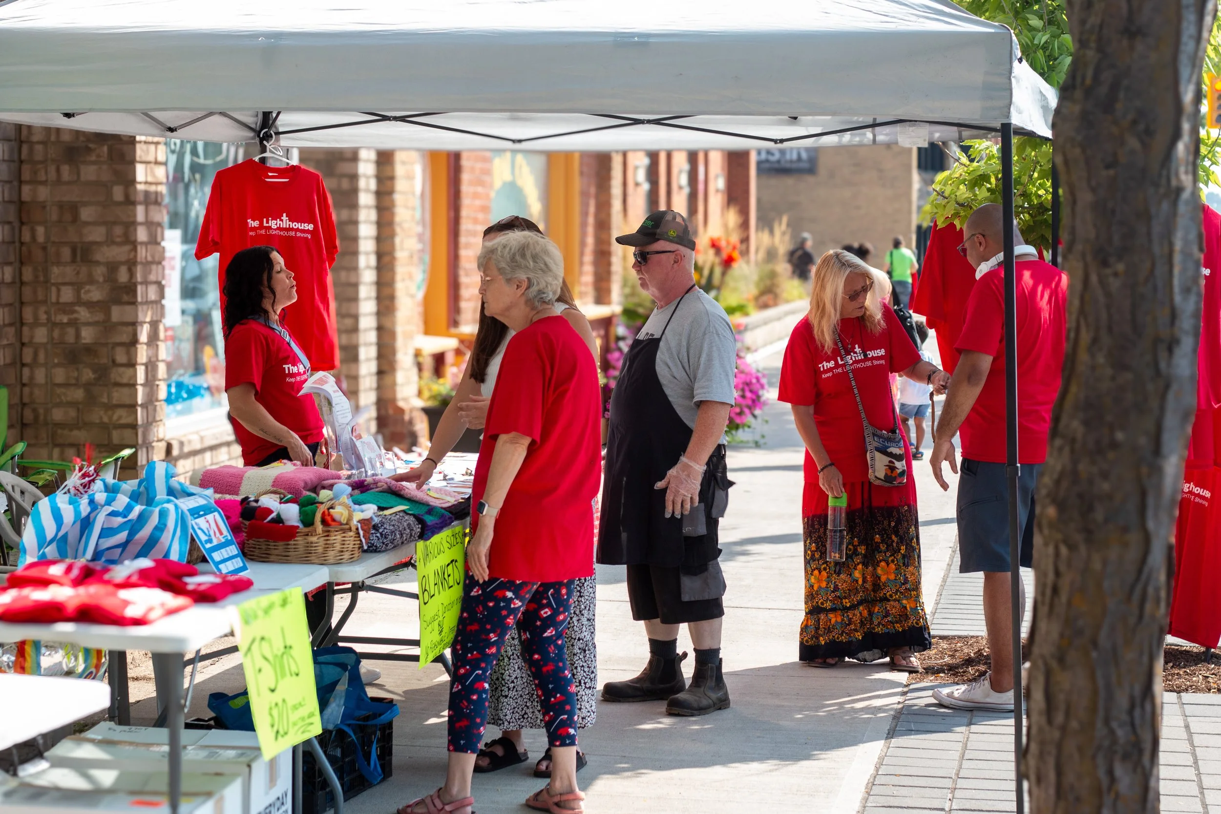 People shopping at an outdoor market stall during daytime. The stall has red T-shirts with 'The Lighthouse' logo and other items like blankets and socks, with bright yellow signs indicating prices.
