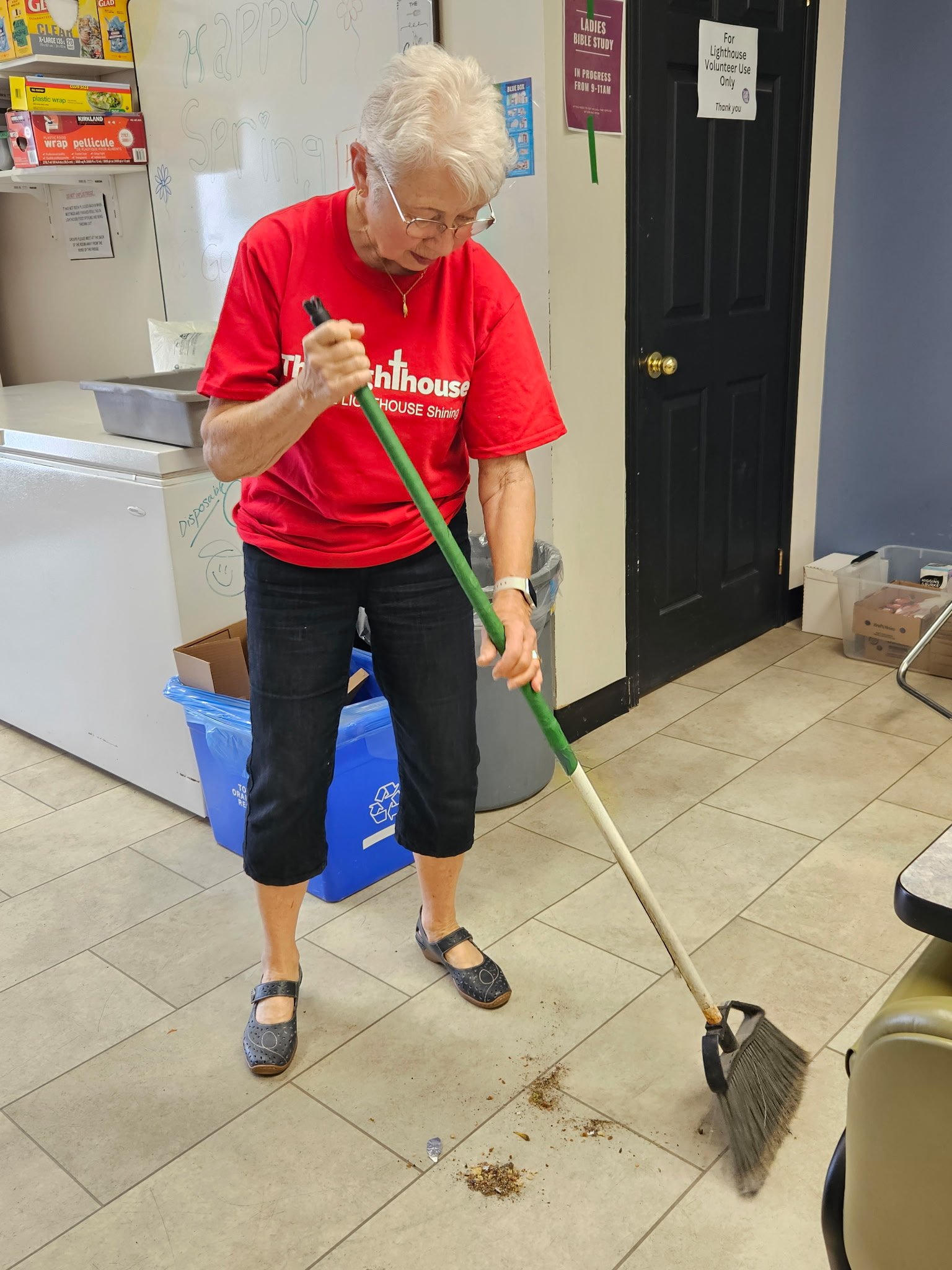 An elderly woman in a red T-shirt and black capri pants sweeping dirt off the floor in an indoor room.
