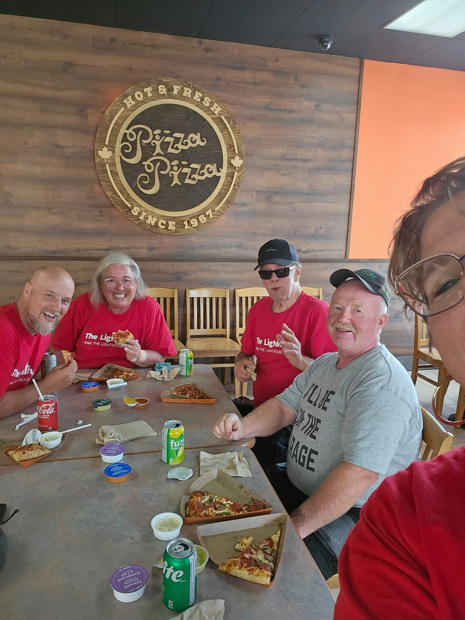 Group of five people sitting at a table enjoying pizza inside a restaurant with a wooden wall and a circular sign that reads 'Pizza Pizza Since 1967' in the background.
