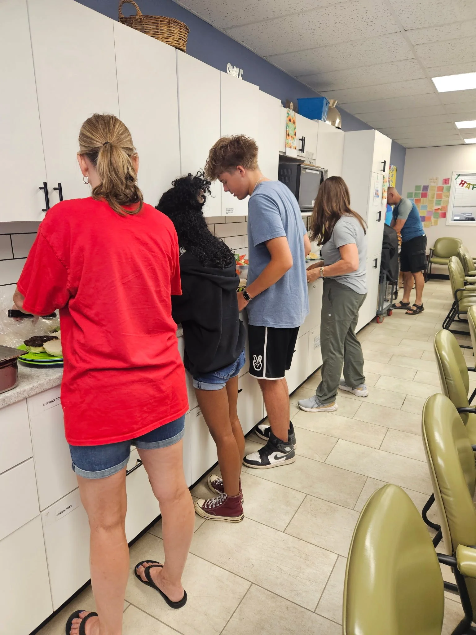 People standing in a line at a kitchen counter in a room with white cabinets and green chairs, preparing or serving food.