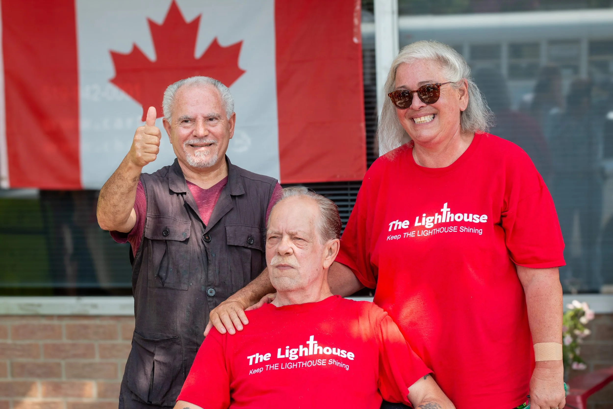 Three seniors standing in front of a Canadian flag, two of them wearing red shirts that say 'The Lighthouse Keep THE LIGHTHOUSE Shining'. One senior is seated, wearing sunglasses and a red shirt, with a woman standing beside him. Another man stands t