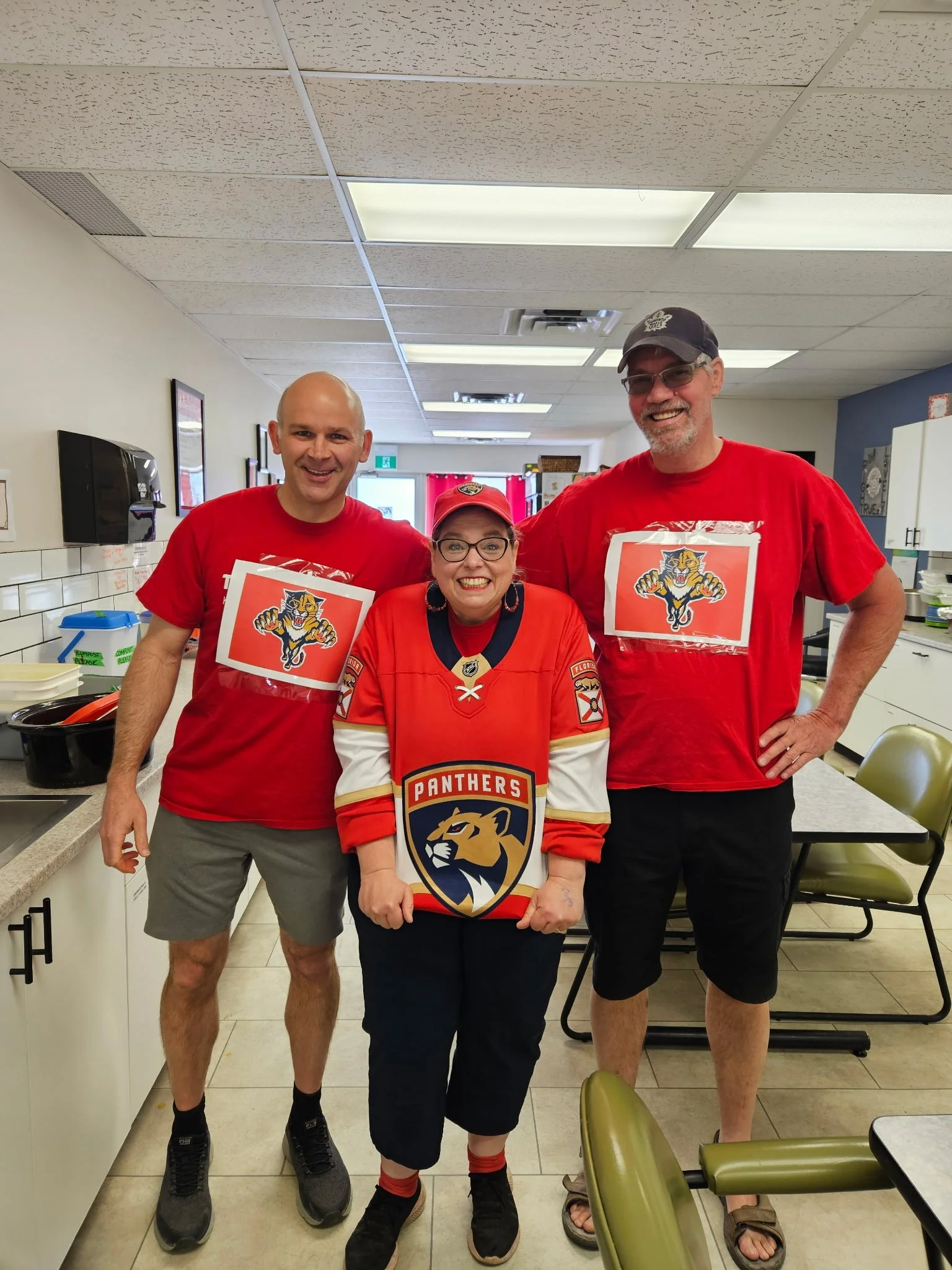 Three people standing in a kitchen, wearing Florida Panthers hockey jerseys and fan gear, smiling at the camera.