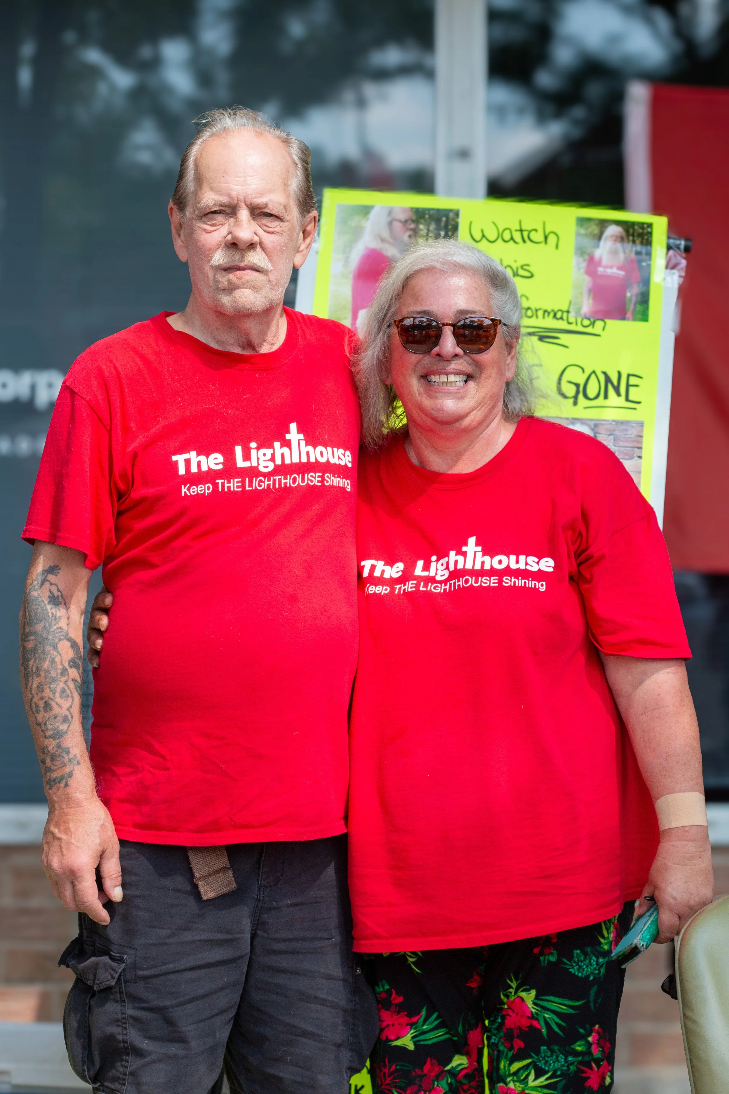 A smiling woman and a serious man standing outdoors, both wearing red T-shirts with 'The Lighthouse Keep THE LIGHTHOUSE Shining' written on them, in front of a yellow sign with text.