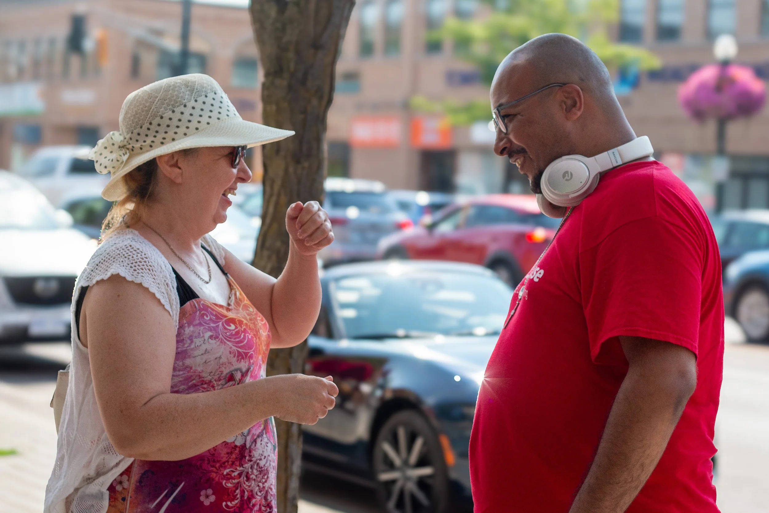 A woman wearing a wide-brimmed hat and glasses smiling and gesturing with her hand while talking to a man in a red t-shirt wearing glasses and headphones around his neck, standing outdoors on a sunny day with cars parked in the background.
