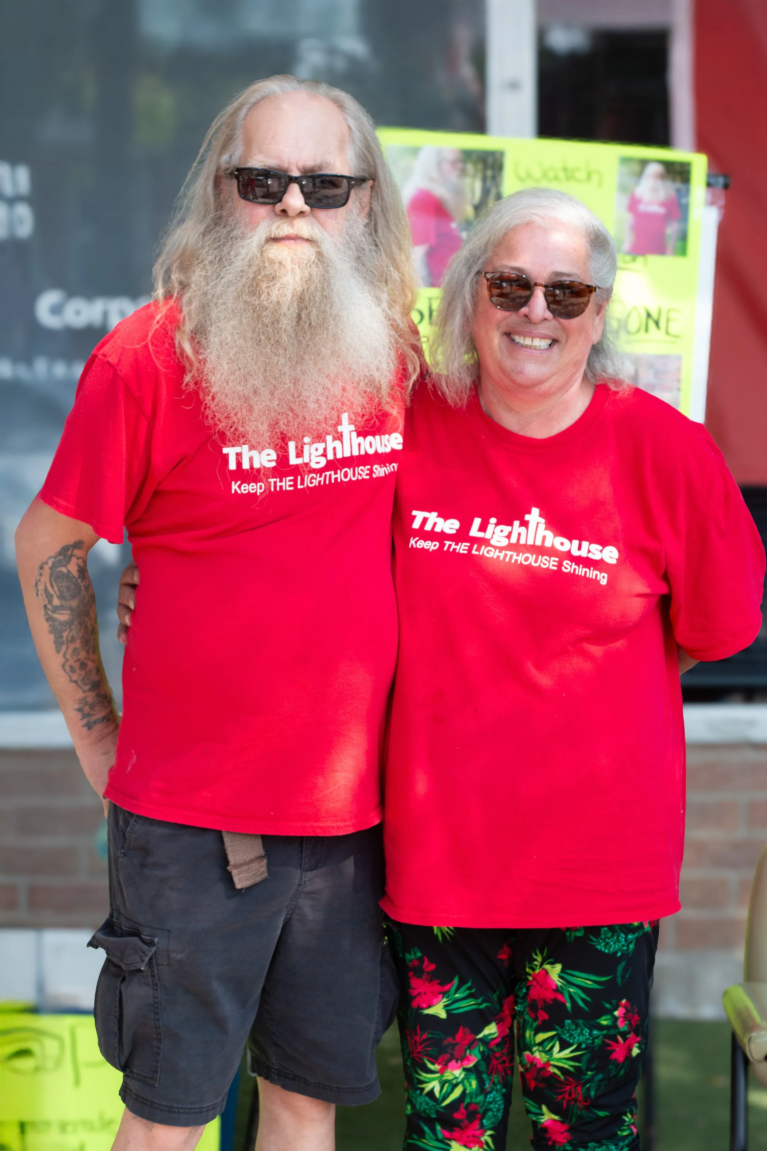 Two people wearing red T-shirts that say 'The Lighthouse Keep THE LIGHTHOUSE Shining' are standing outdoors. The man has long hair and a beard, sunglasses, and tattoos on his arm. The woman has gray hair, sunglasses, and floral-patterned pants. They 