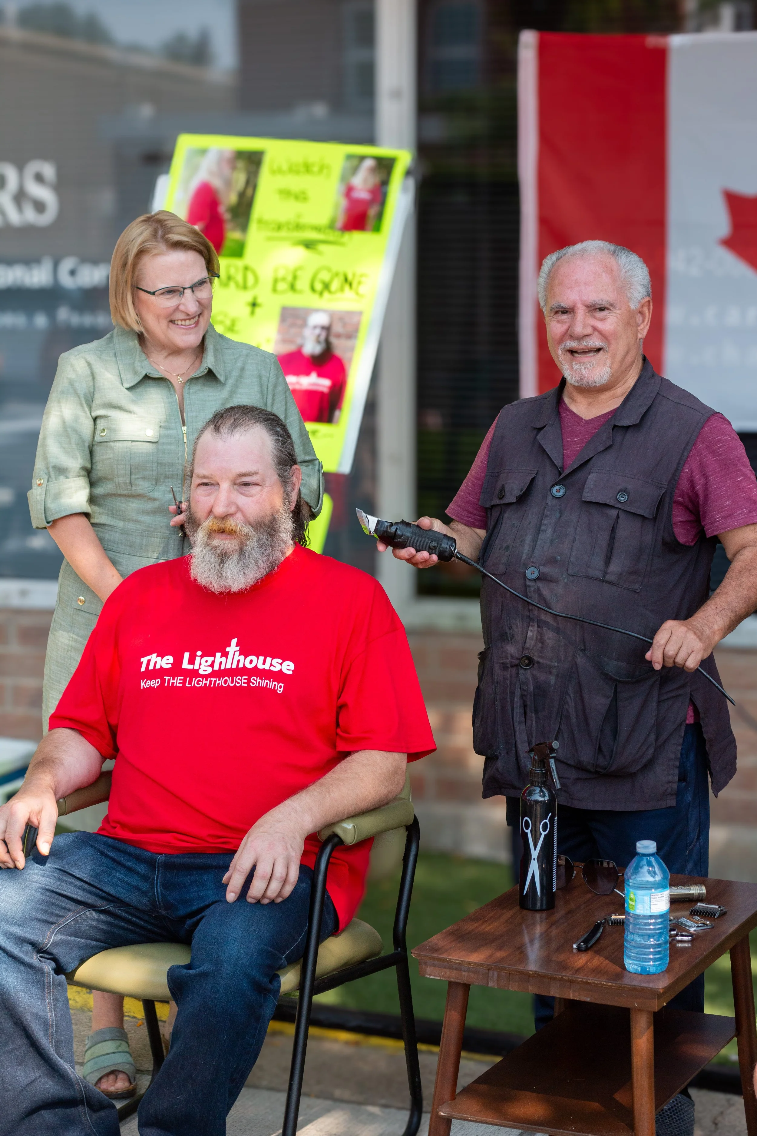 A man with a beard and red T-shirt sitting in a chair while another man with gray hair and a beard styles his hair outdoors. A woman stand nearby smiling, and a table holds barber tools and water bottle.