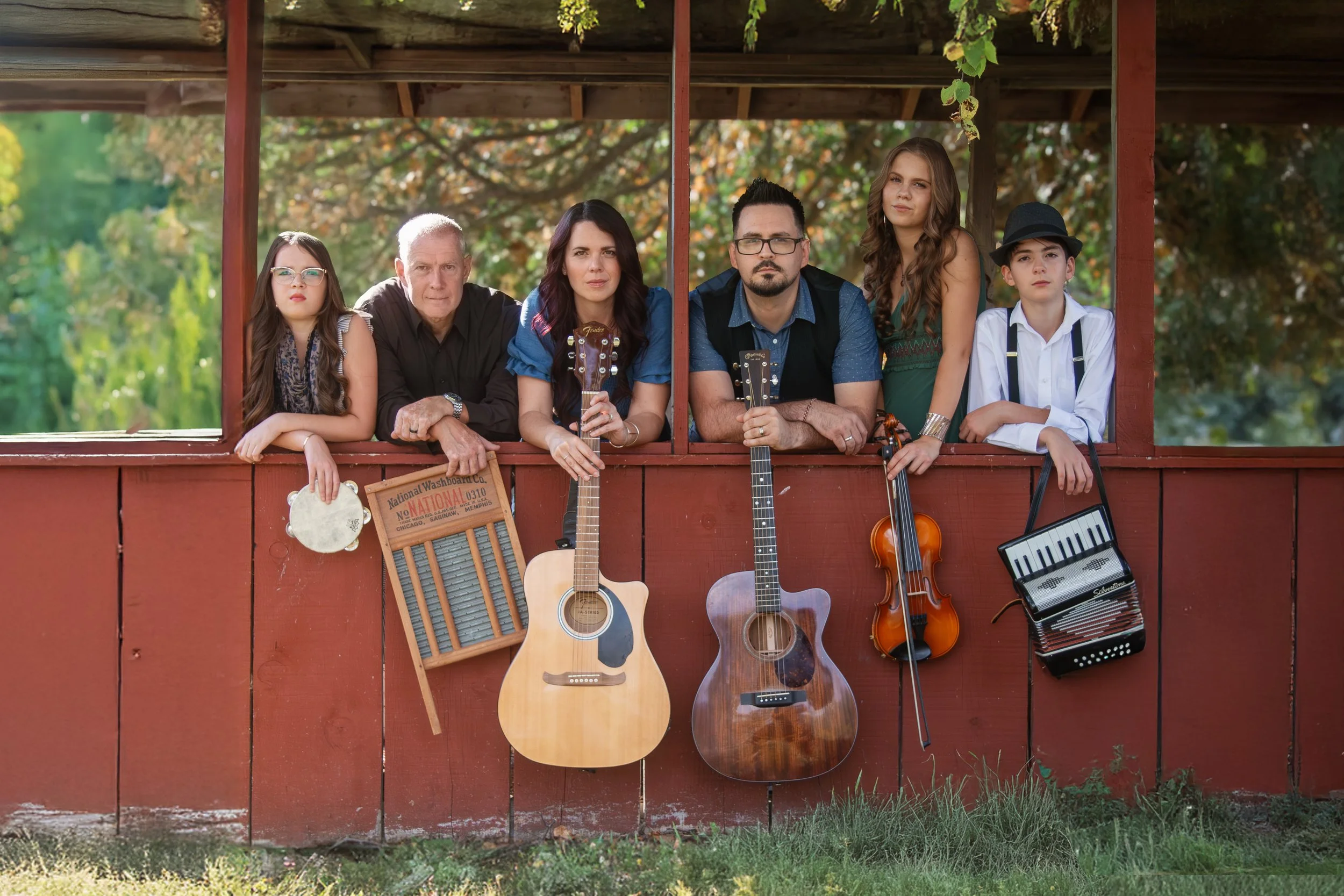 Family group of six people with musical instruments leaning on a red wooden fence outdoors, with trees in the background and sunlight filtering through. They have guitars, a violin, a tambourine, a washboard, and an accordion.