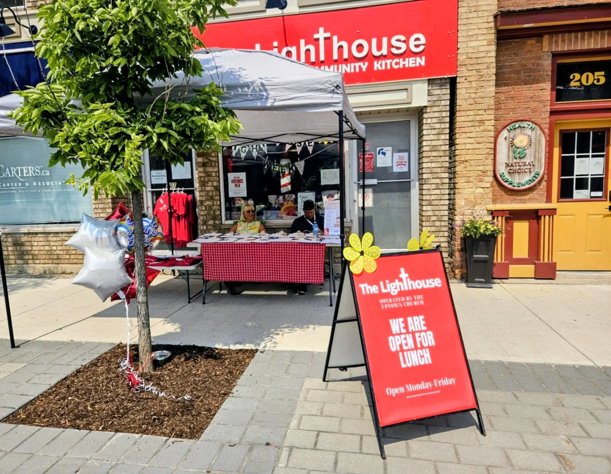 Street scene with a red signboard for The Lighthouse Community Kitchen, an outdoor tent with two people sitting at a table, and a sidewalk sign indicating lunch is available Monday through Friday.