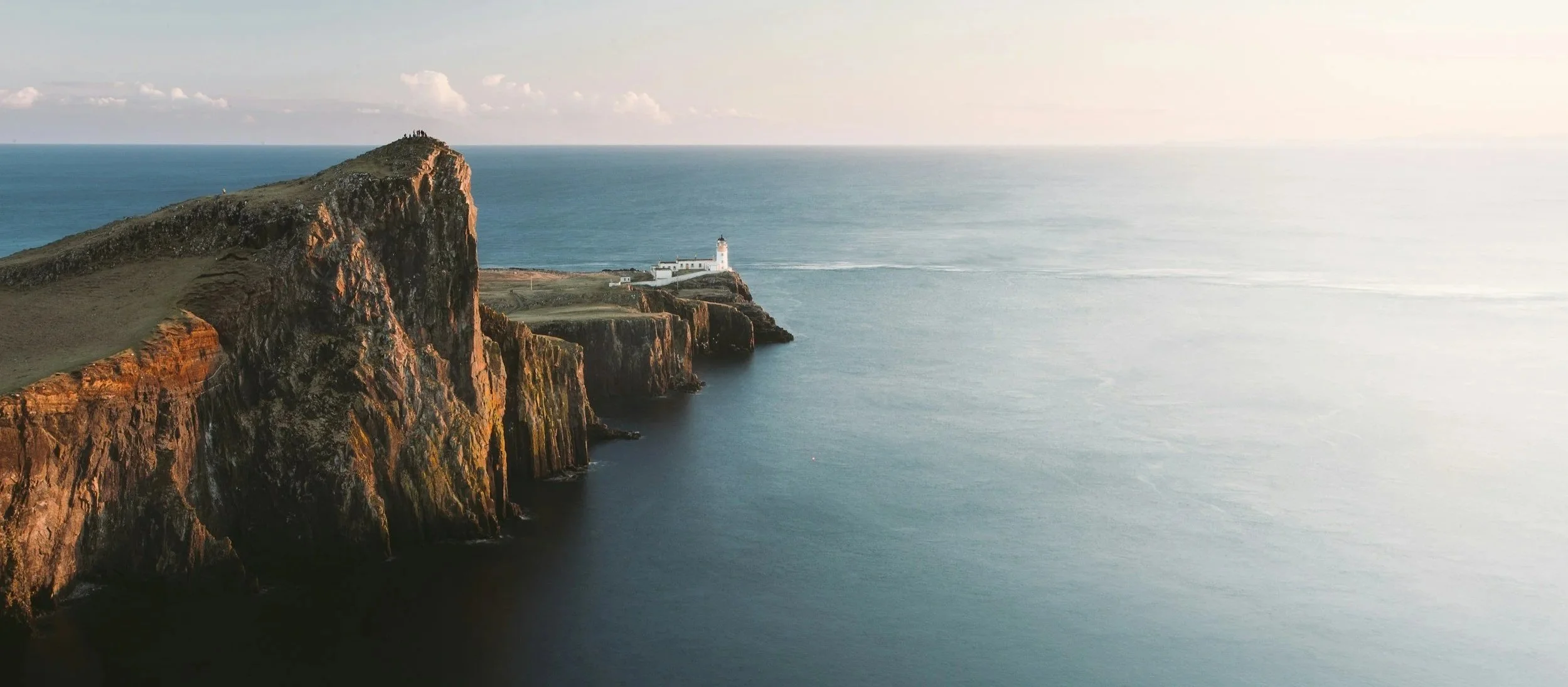 Cliffs overlooking the ocean with a lighthouse on the edge of a headland and a building nearby, under a partly cloudy sky.